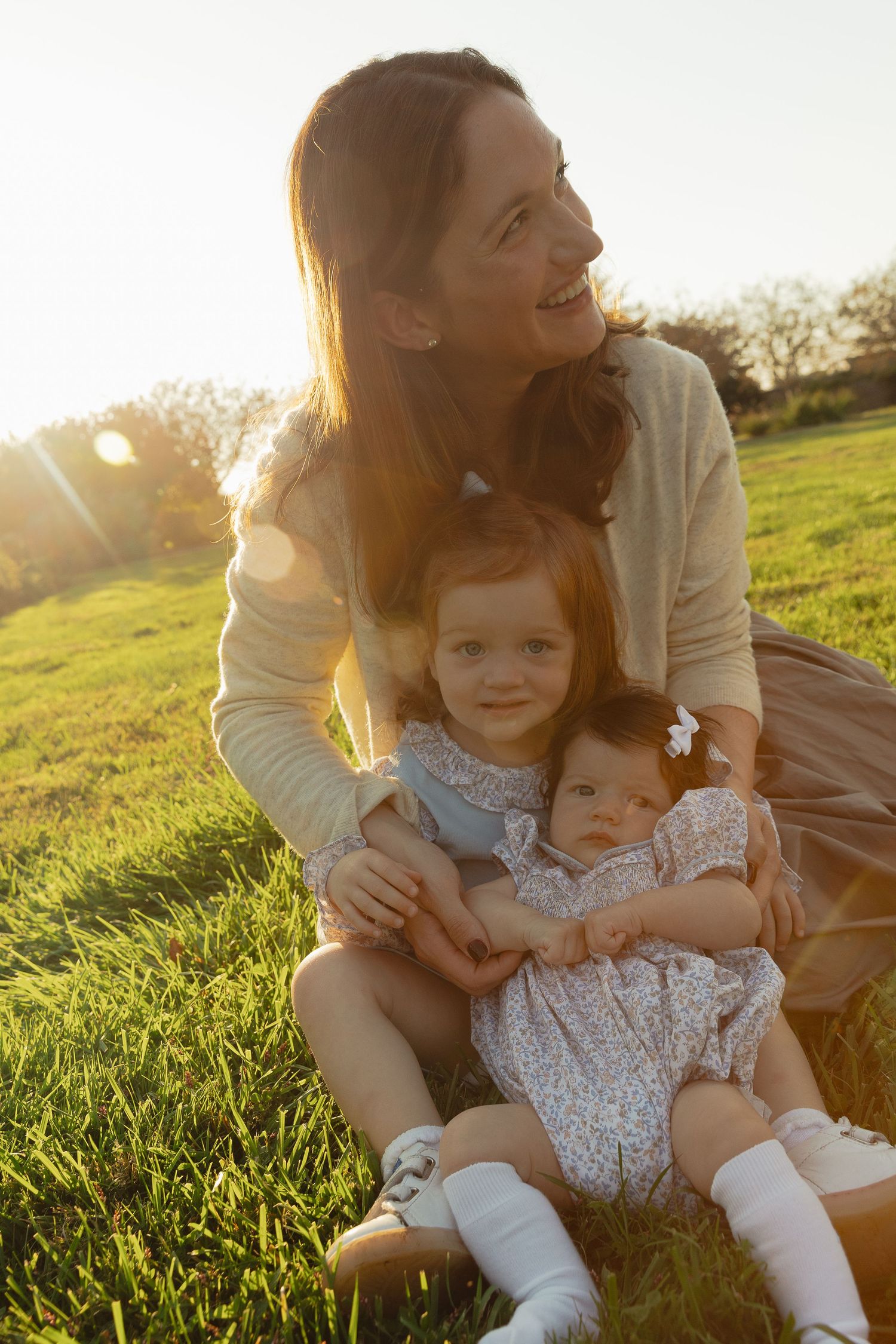 A smiling mother sits on green grass with her two young daughters in matching floral dresses during golden hour sunset.
