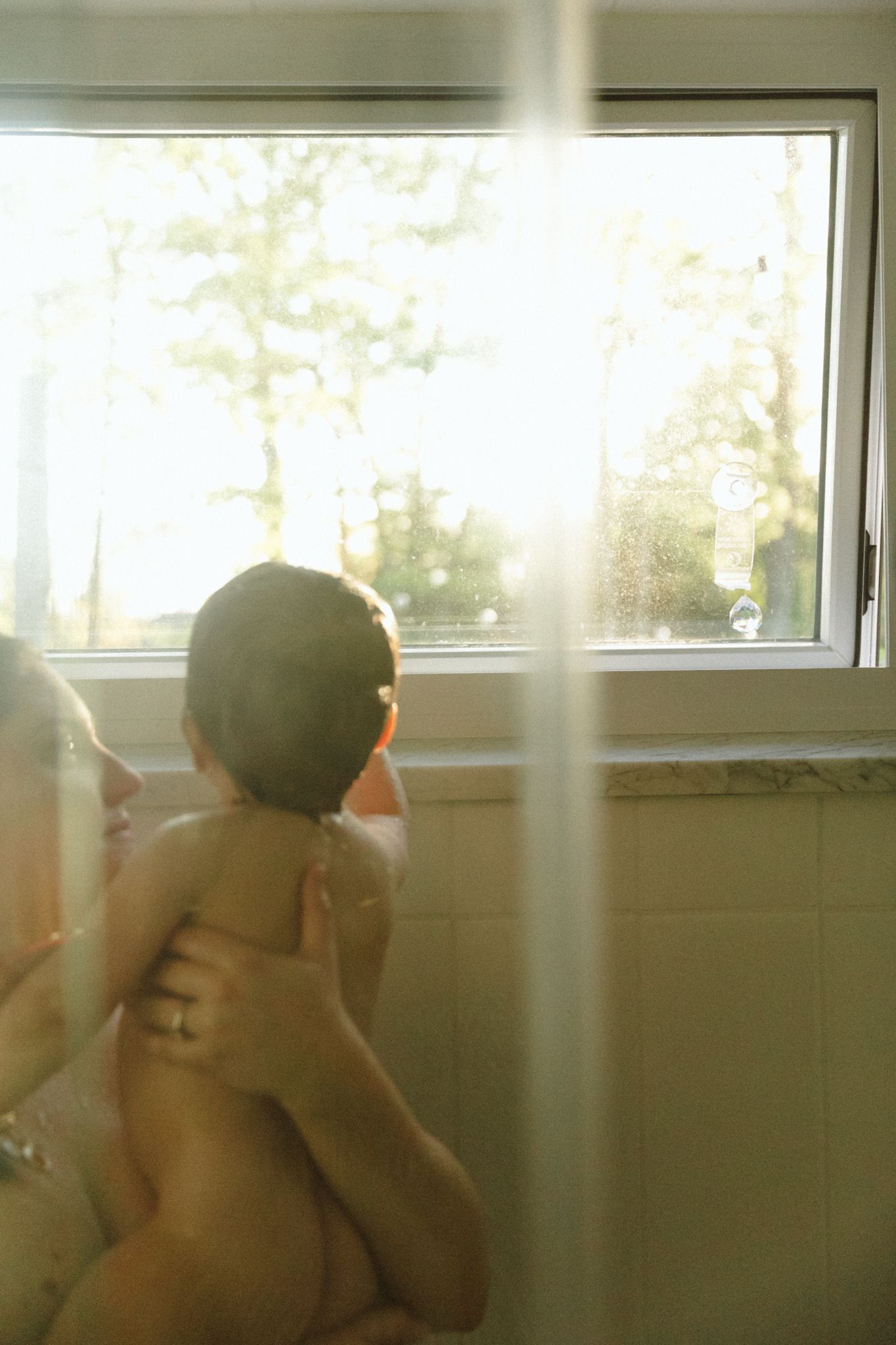 Parent bathing baby near sunlit bathroom window with warm natural light and trees visible outside.