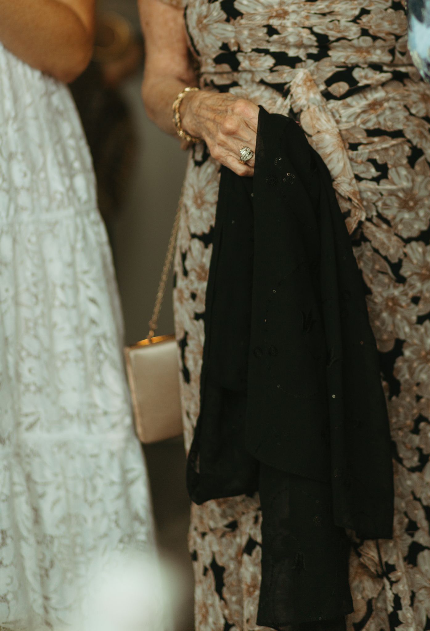 Elderly woman in floral lace dress holding black fabric and gold rings, standing beside woman in white lace dress.