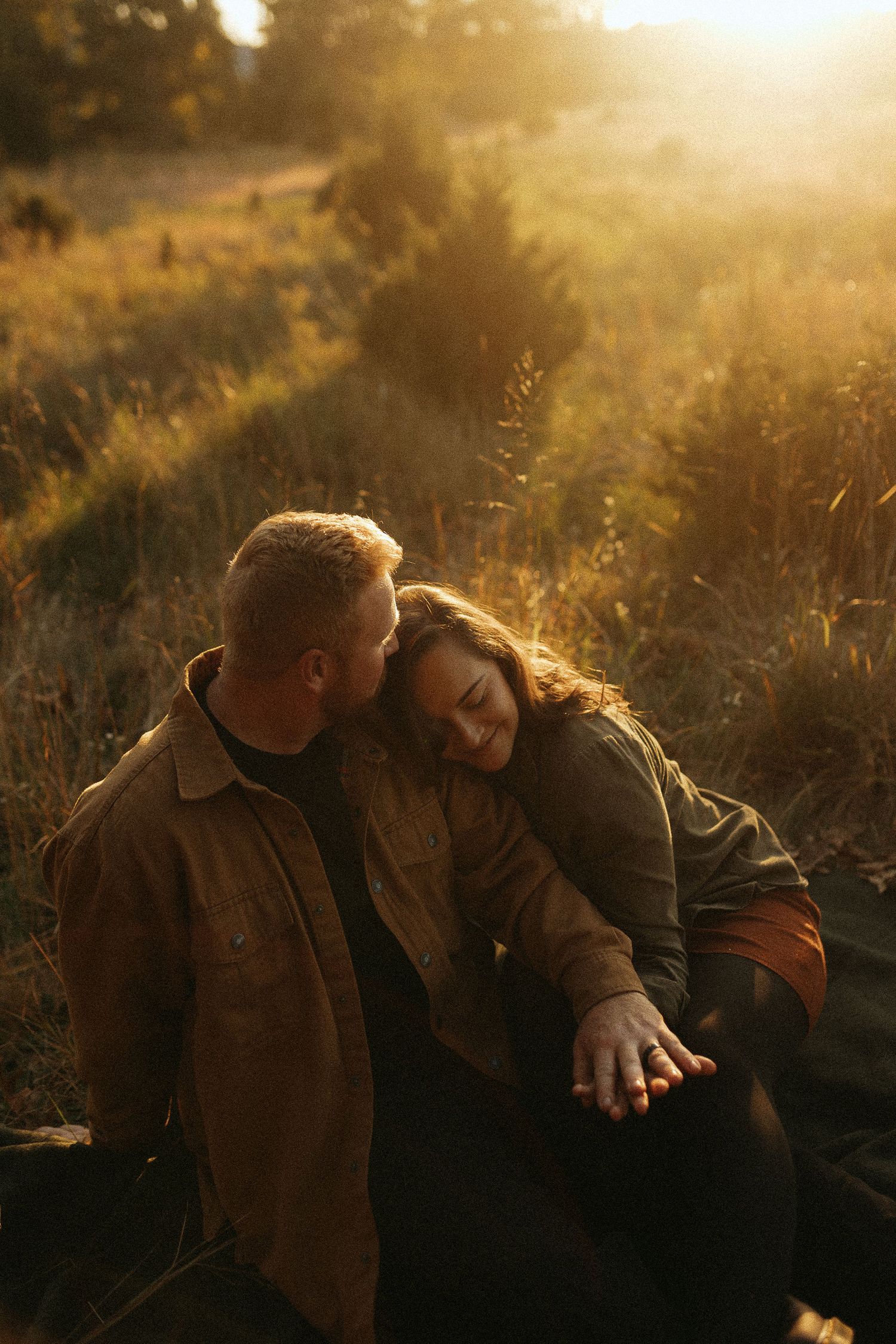 Couple sitting in golden sunset field, woman resting head on man's shoulder, holding hands, warm autumn light.