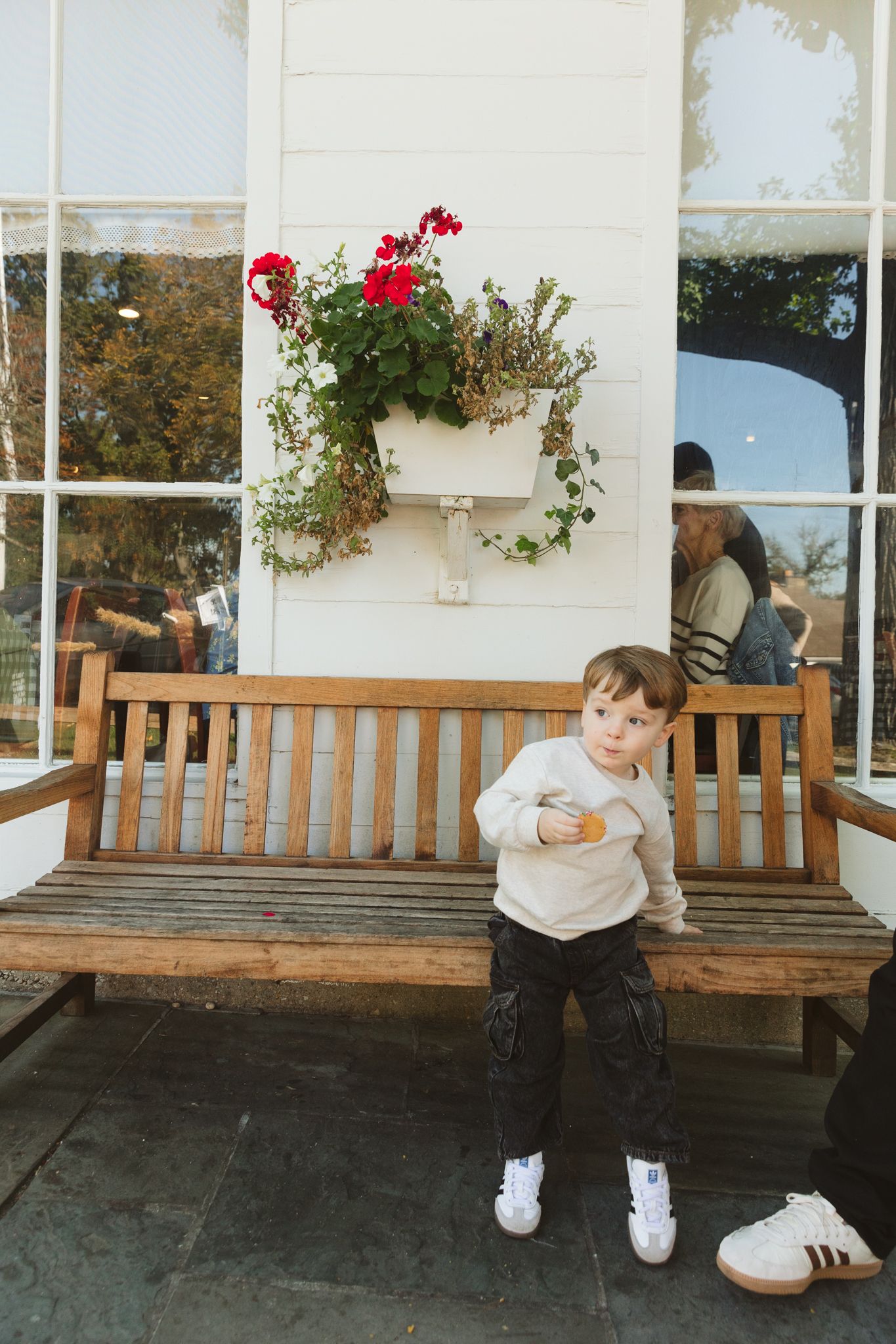 A young toddler in a cream sweater and black pants sits on a wooden bench outside a white building with red flowers.