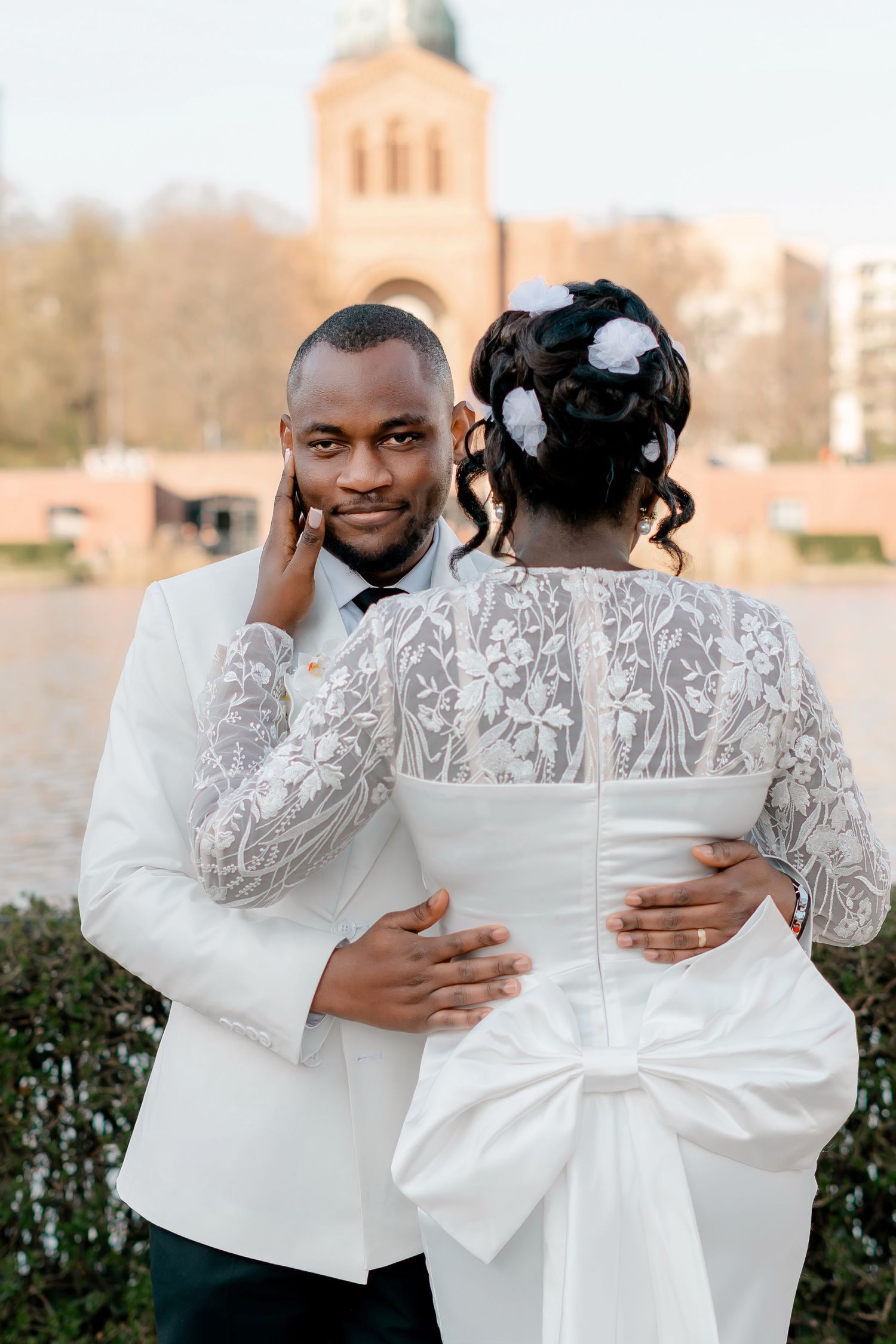 Black groom in white tuxedo embracing bride in lace wedding dress with a church in the background.