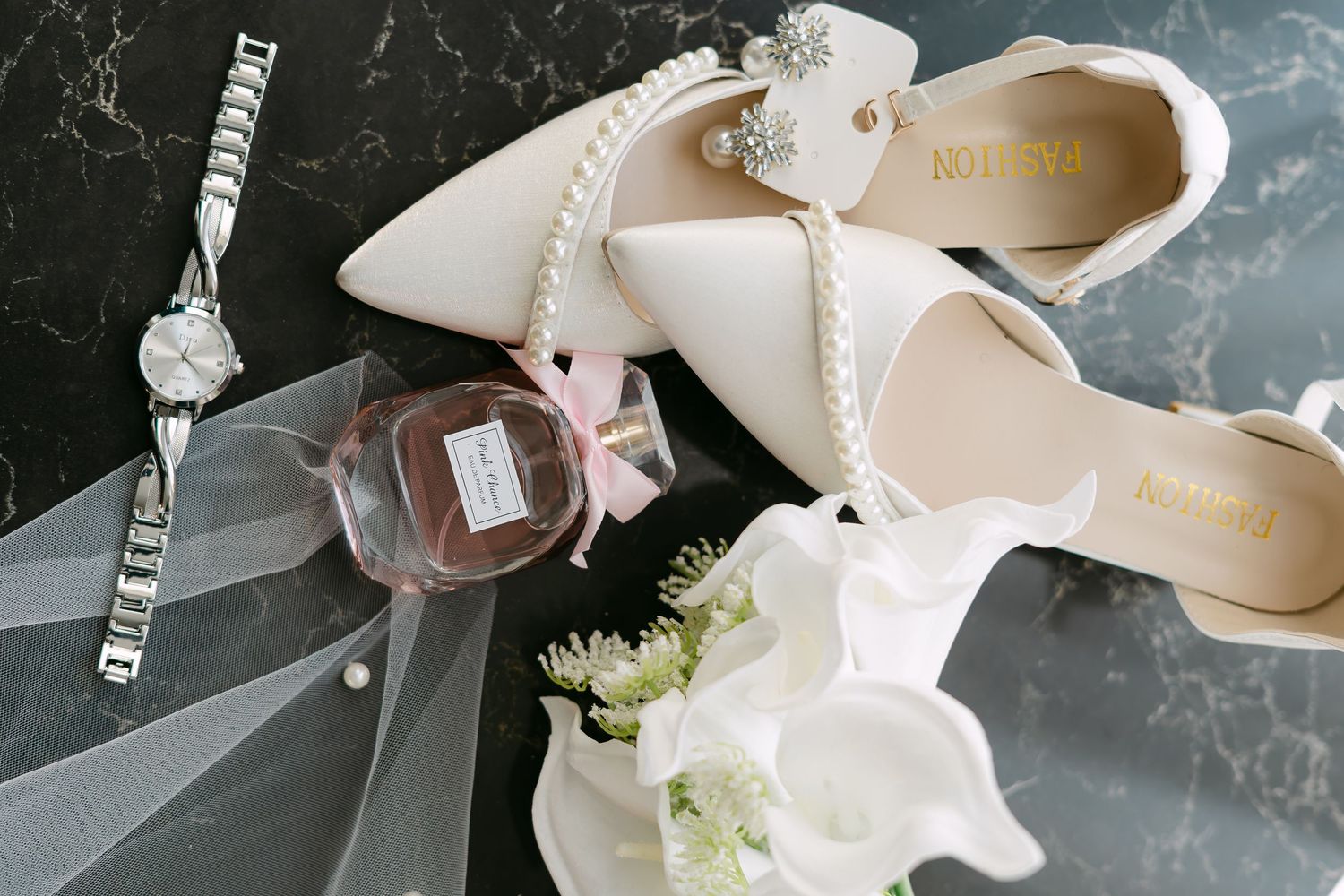 Bridal accessories flat lay with pearl heels, perfume, watch, and white floral bouquet.