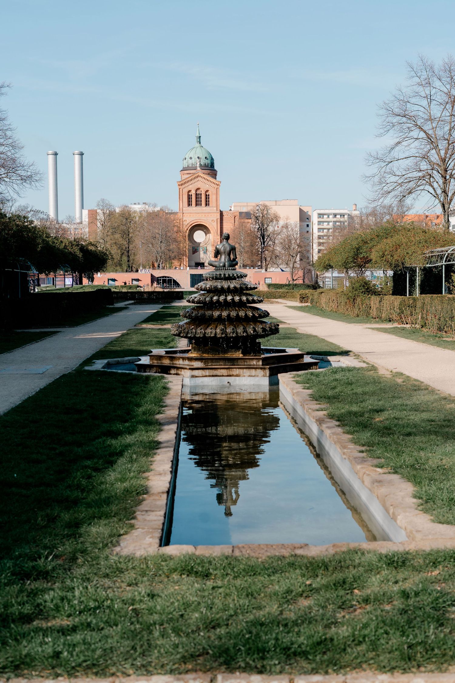 A ornate tiered fountain reflects in a rectangular pool in a park with a historic red brick church in the background.