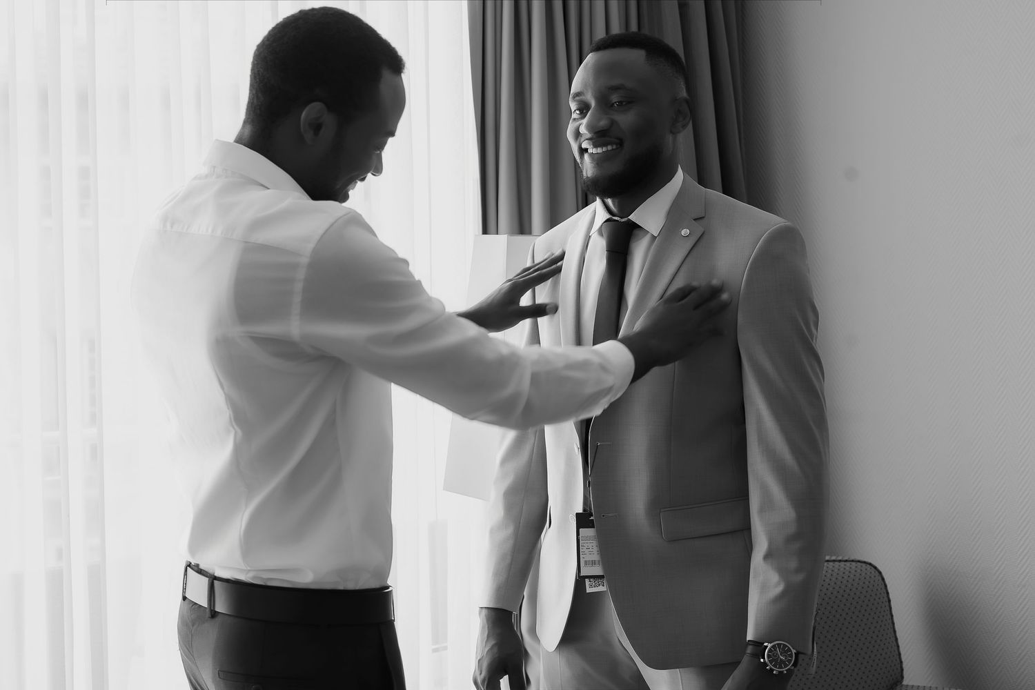 Black and white photo of two men in suits, one adjusting the other's tie, preparing for a formal event.