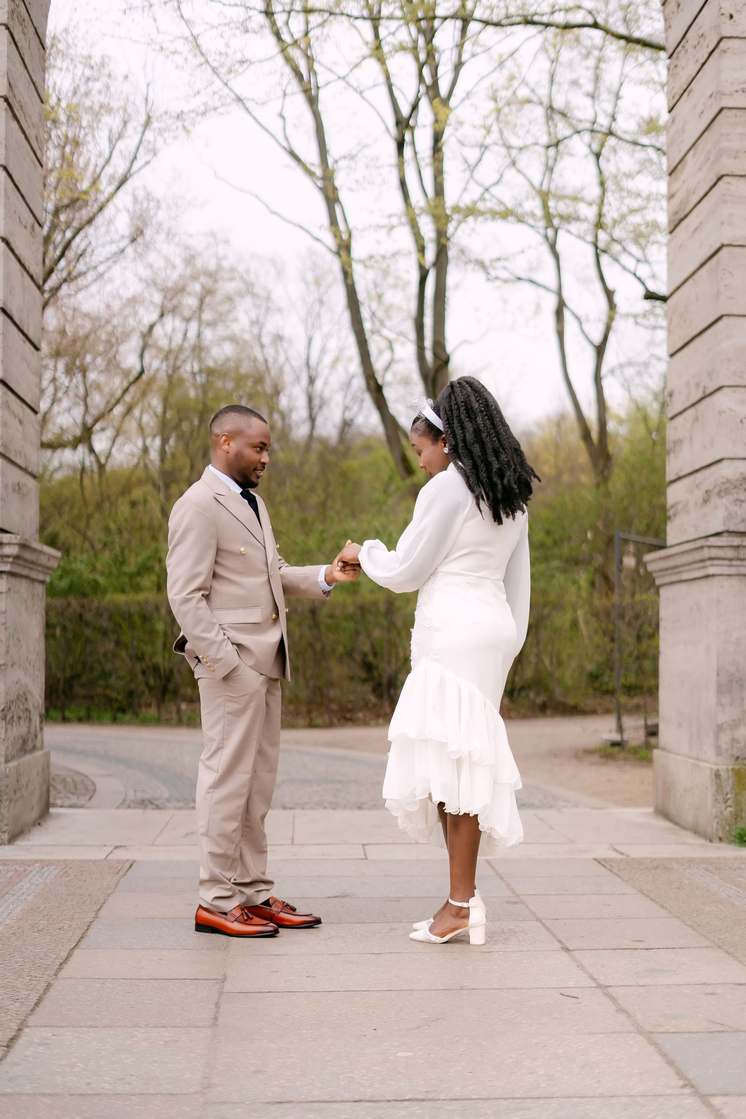 A bride and groom hold hands while standing between two stone columns in a park setting in spring.