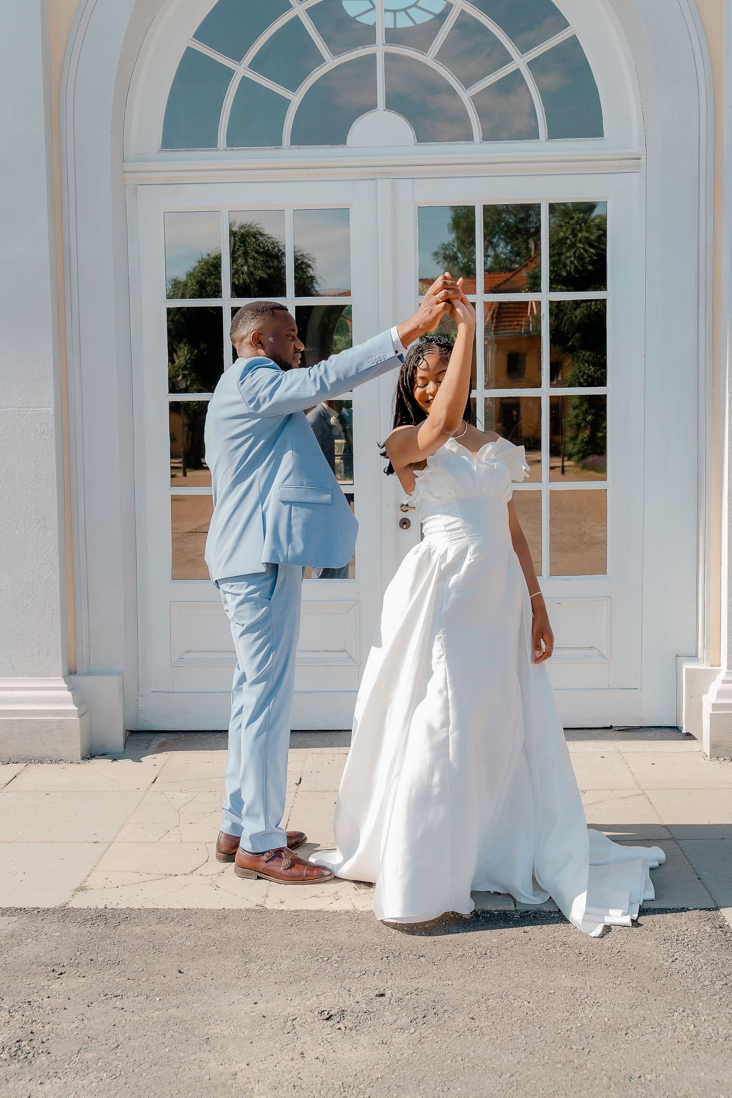 A groom in a light blue suit twirls his bride in a white wedding gown outside an elegant white building.