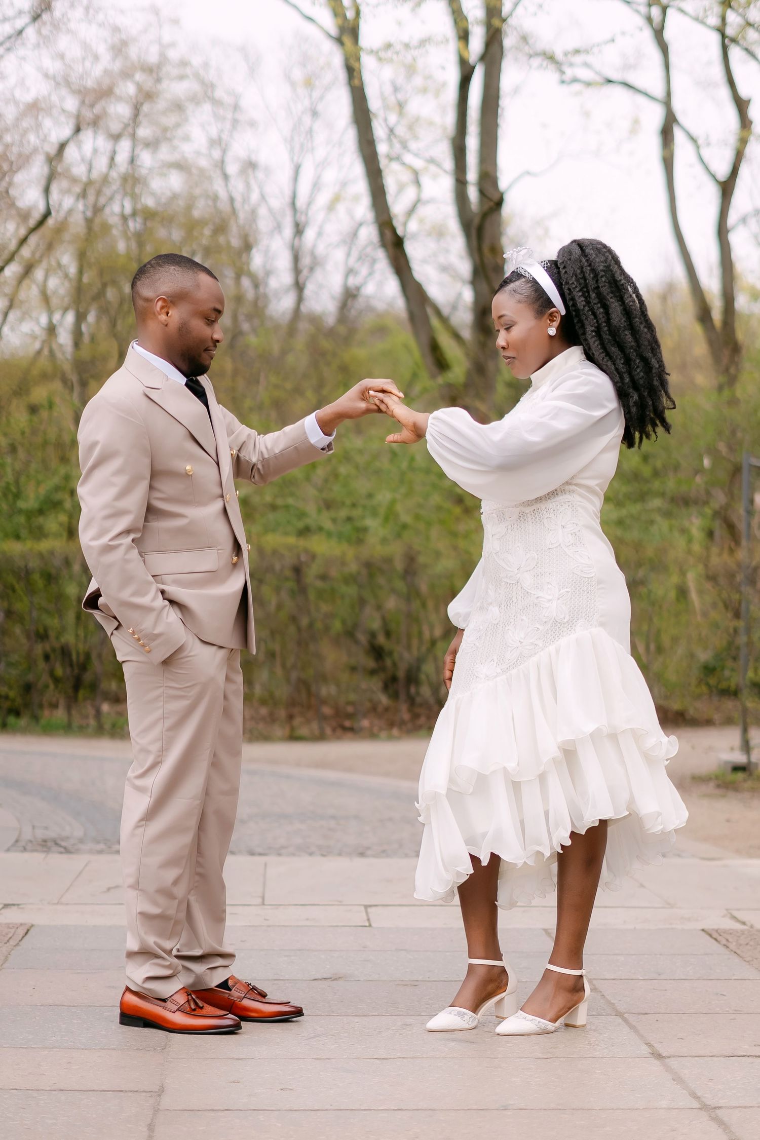 Black wedding couple holding hands and dancing outdoors on stone path surrounded by spring trees.