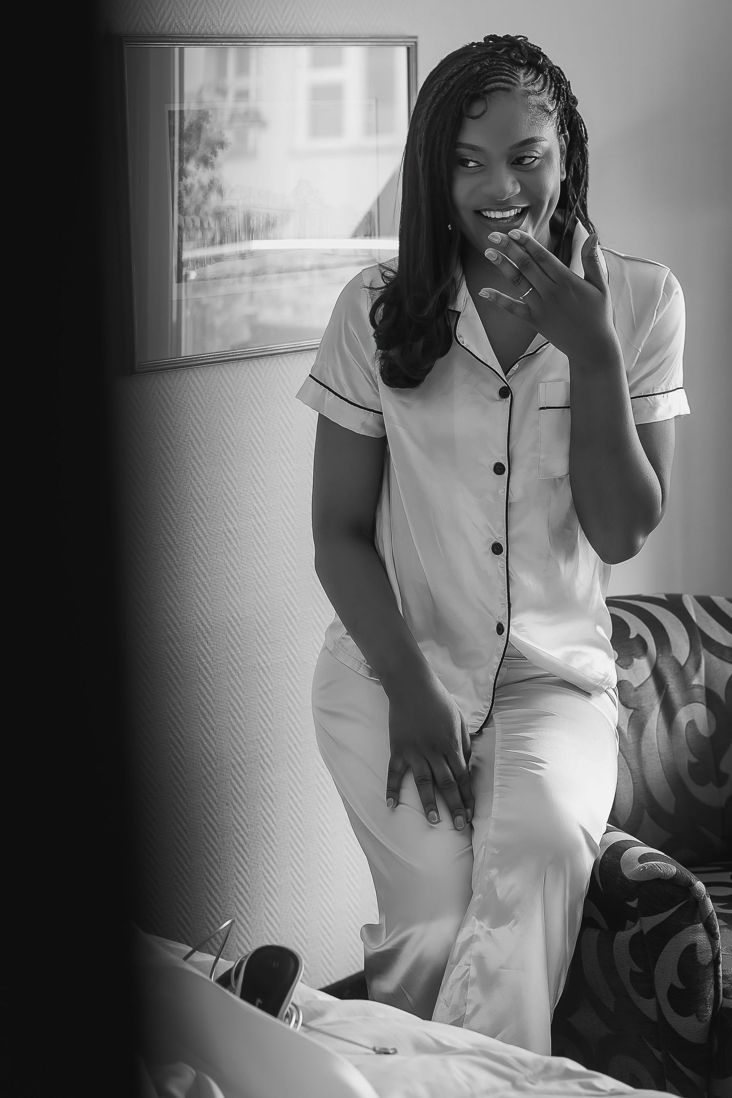 Black and white photo of a smiling young woman in pajamas sitting on a bed in a cozy bedroom.