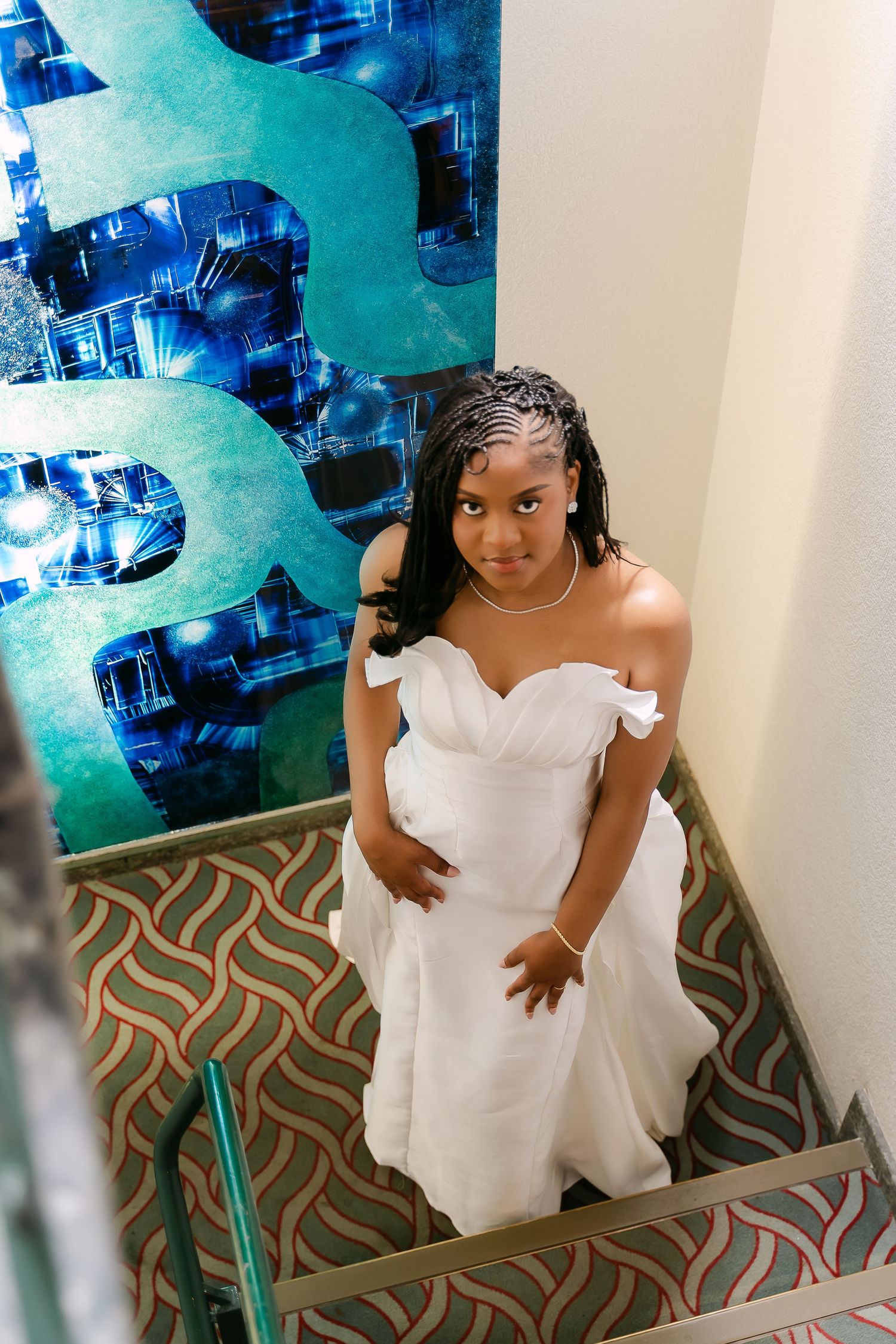 Young bride in white off-shoulder gown ascending stairs, braided hair, pearl necklace, beside teal decorative wall art.