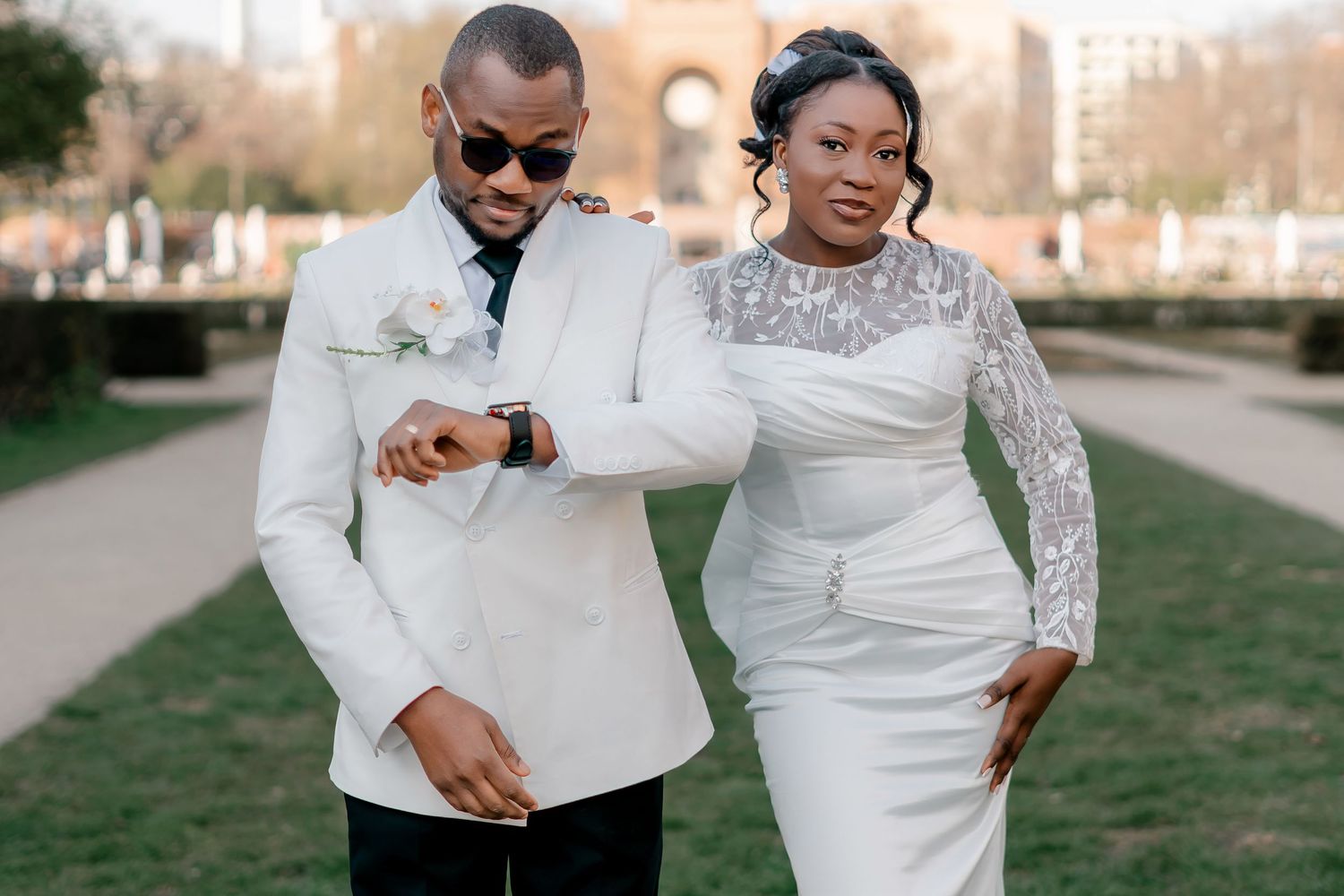 Stylish groom in white suit checking watch while walking with bride in lace wedding dress.