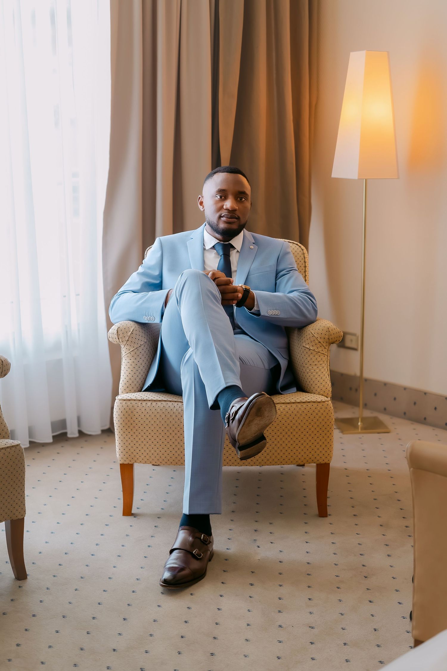 A well-dressed Black man in a light blue suit sits confidently in an armchair, adjusting his tie in an elegant hotel room.