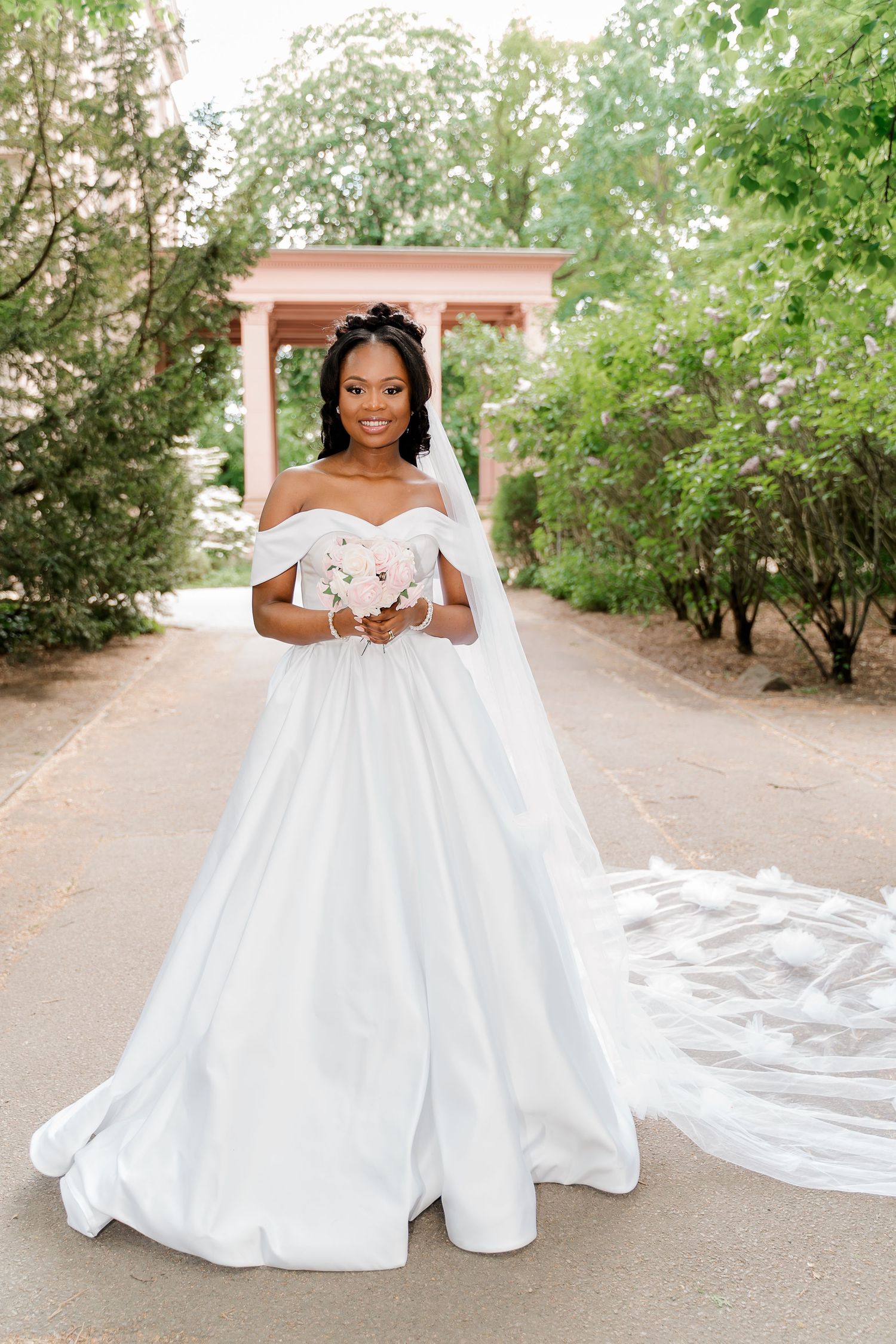 Bride in off-shoulder white ball gown with veil holds bouquet in lush garden setting.