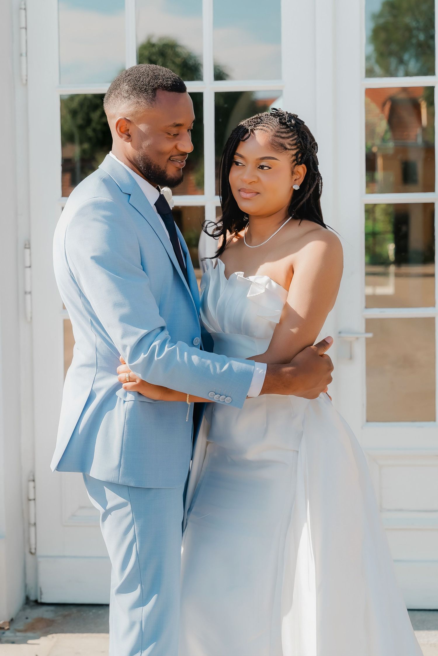 A Black couple in wedding attire, groom in light blue suit and bride in white dress, embracing outdoors.