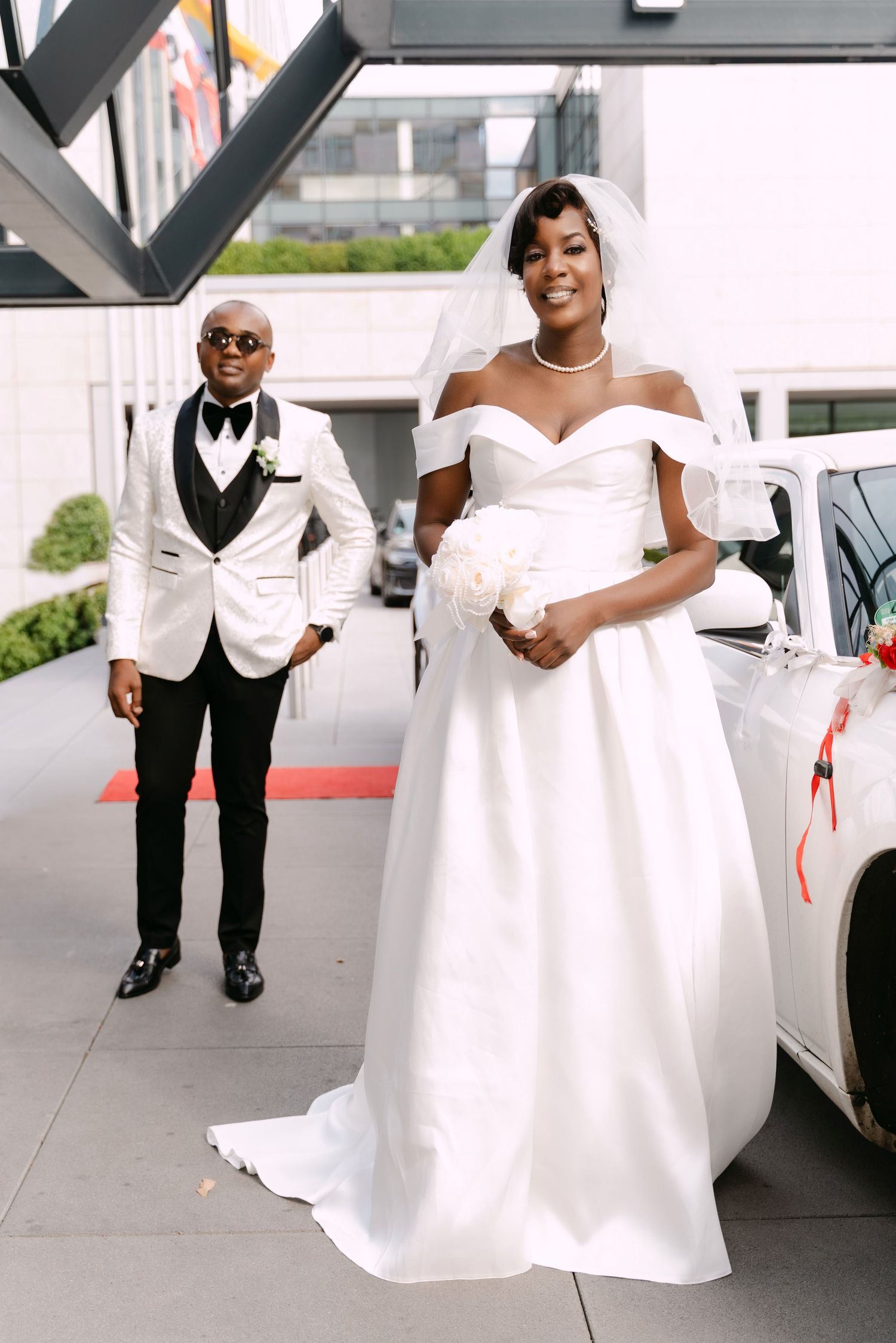 Elegant couple posing outside venue, groom in white tuxedo and bride in off-shoulder wedding gown.