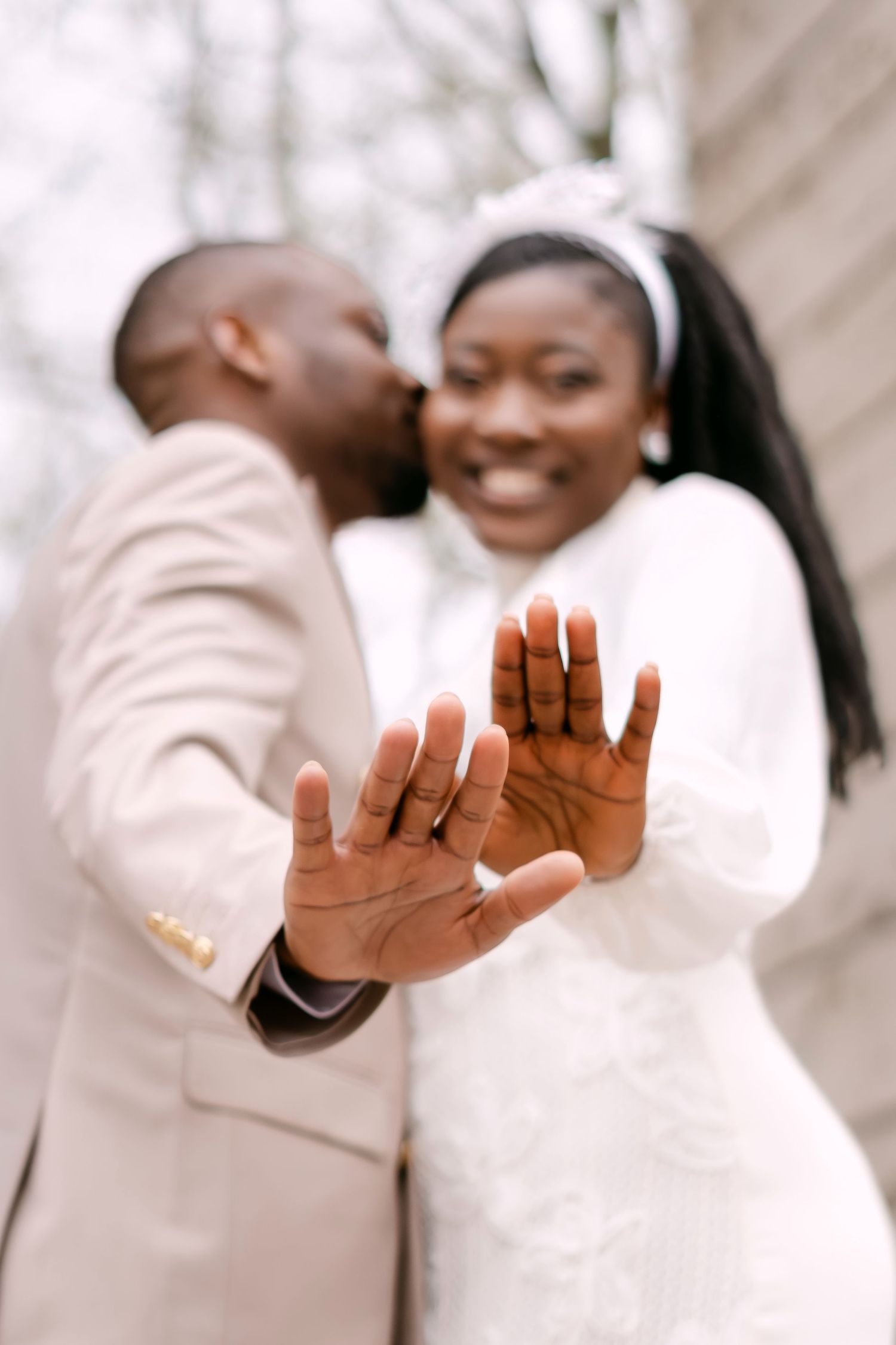 Newlywed Black couple displaying wedding rings to camera, groom kissing bride who wears white headband.