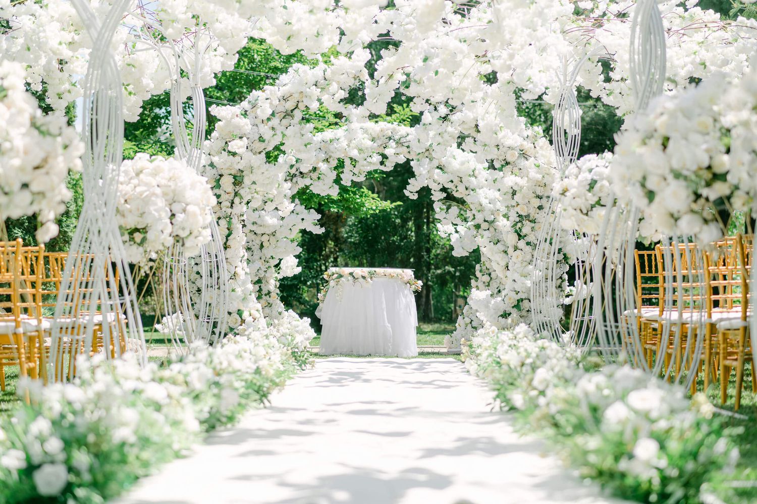 Elegant outdoor wedding ceremony aisle with white floral arches, gold chairs, and lush greenery creating a romantic garden setting.
