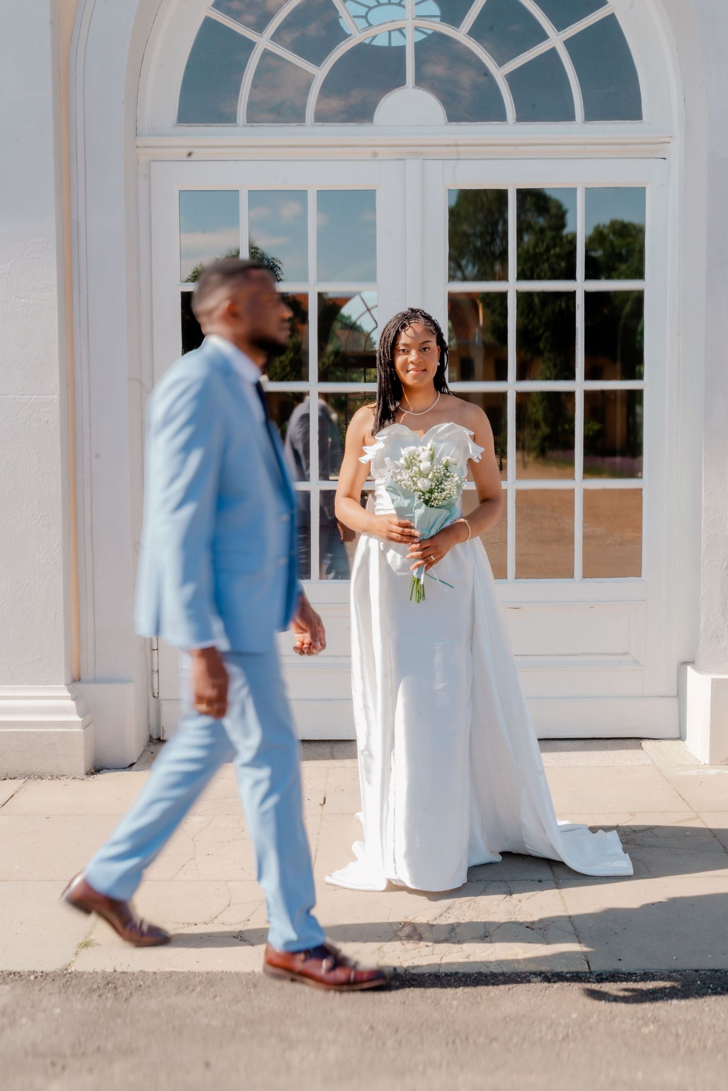 Bride in white gown holds bouquet as groom in light blue suit walks toward her outside elegant white building.