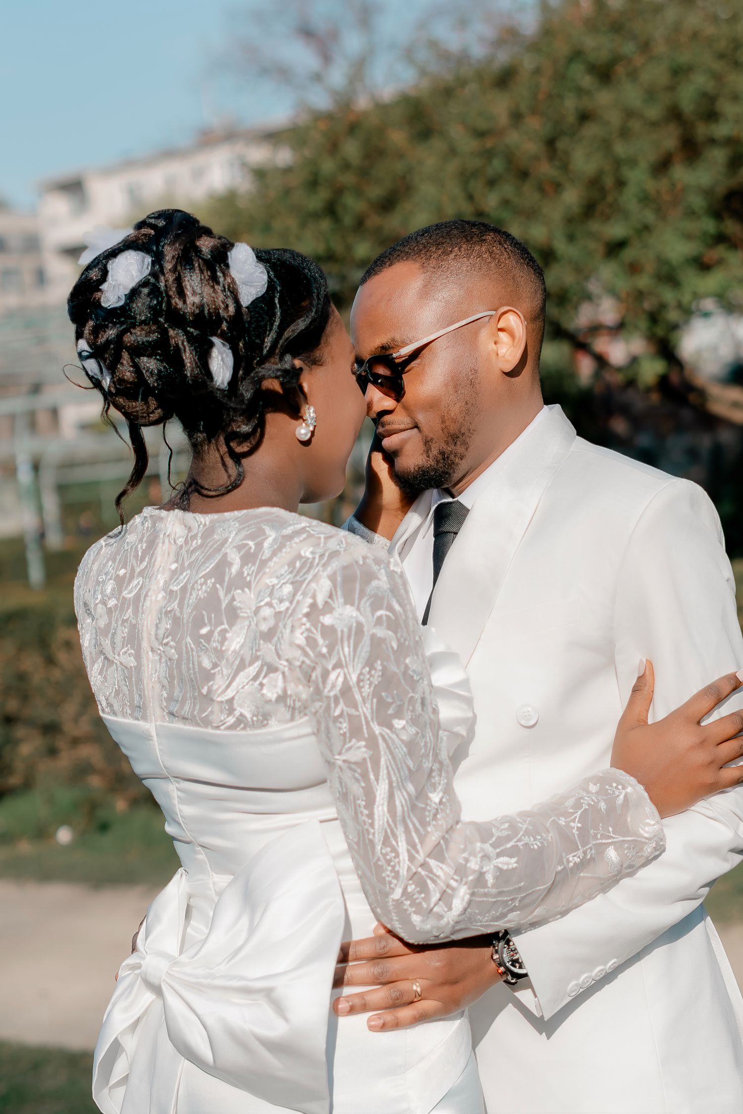 Groom in white suit embracing bride in lace wedding dress outdoors in romantic moment.