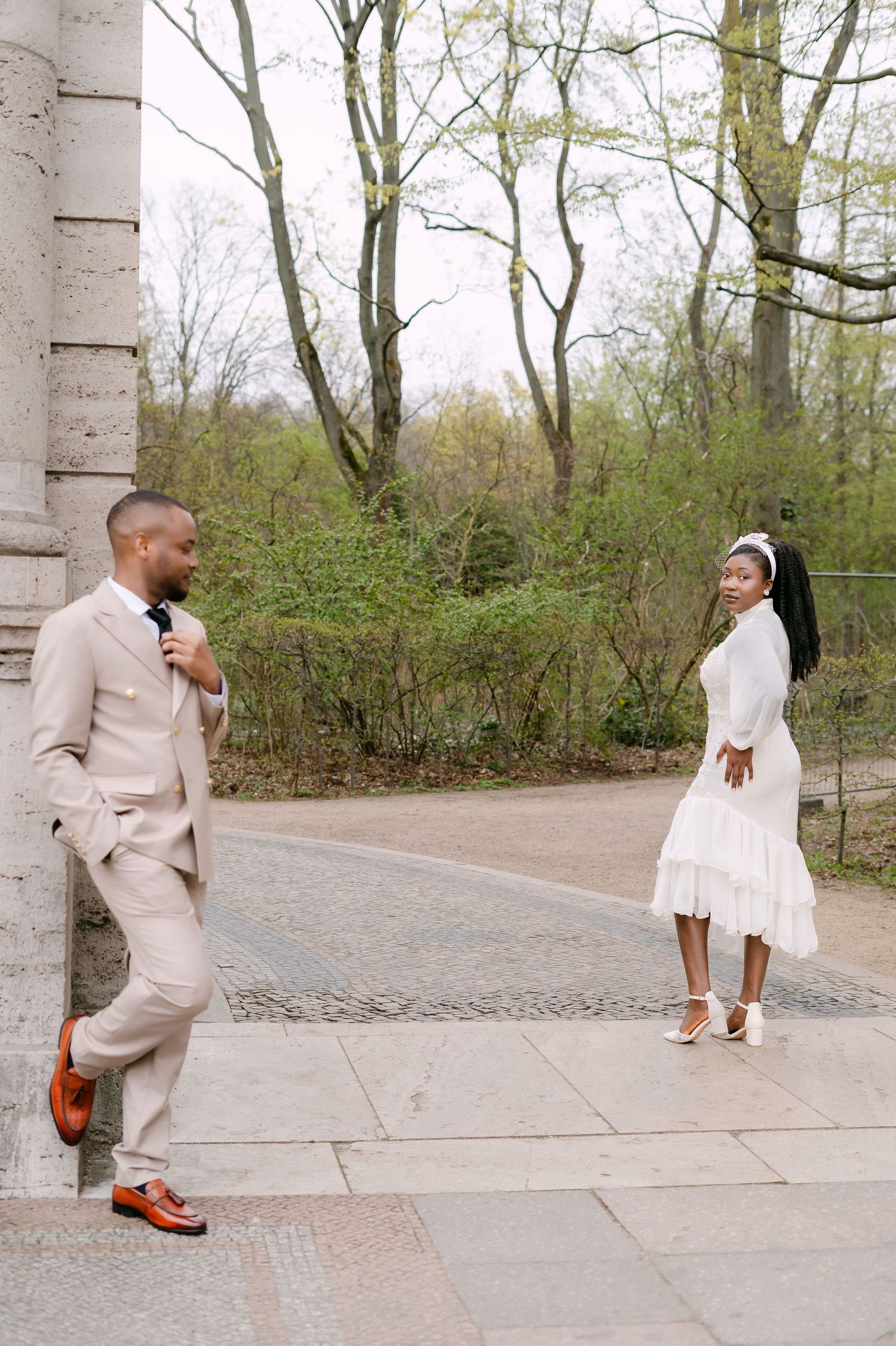 A groom in a beige suit leans on a stone pillar smiling at his bride in a white midi dress outdoors.