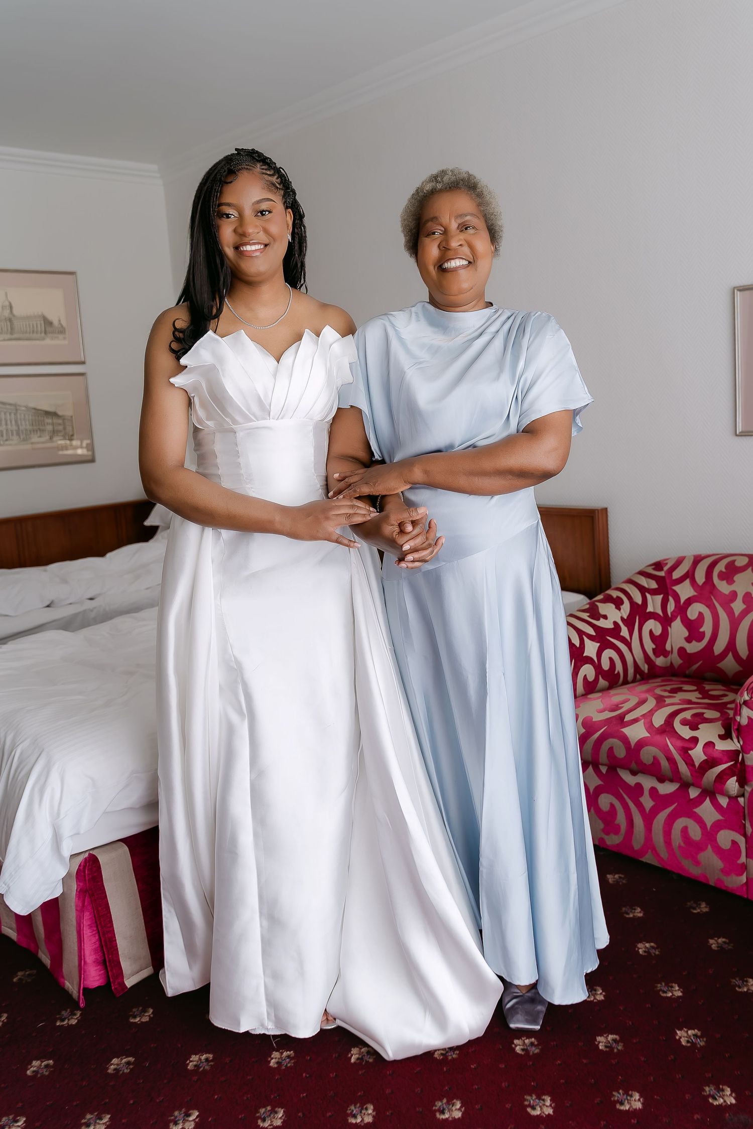 Bride in white gown and older woman in light blue dress smiling together in a hotel room.