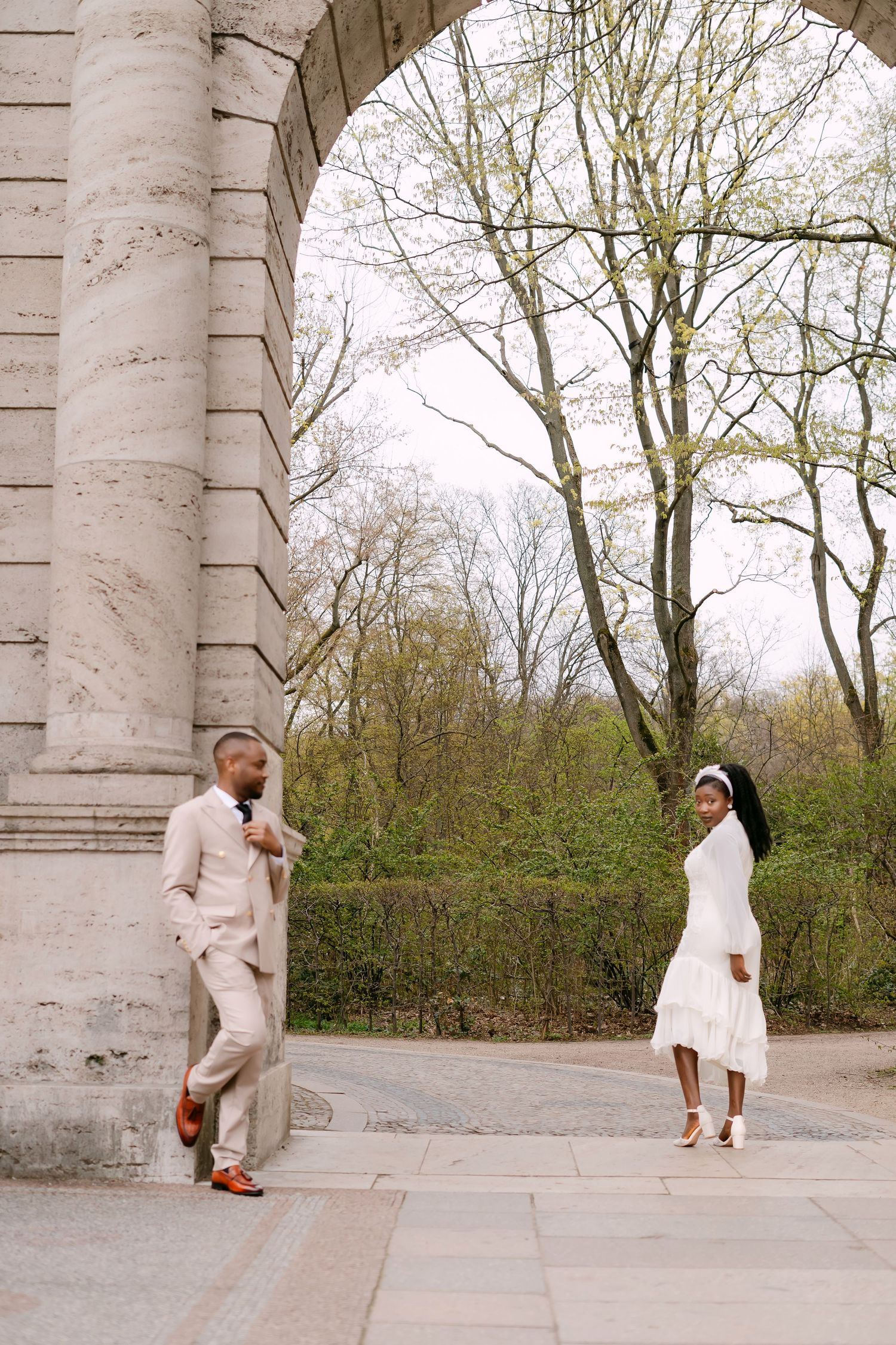 A stylish couple in wedding attire pose under a large stone arch surrounded by early spring trees.