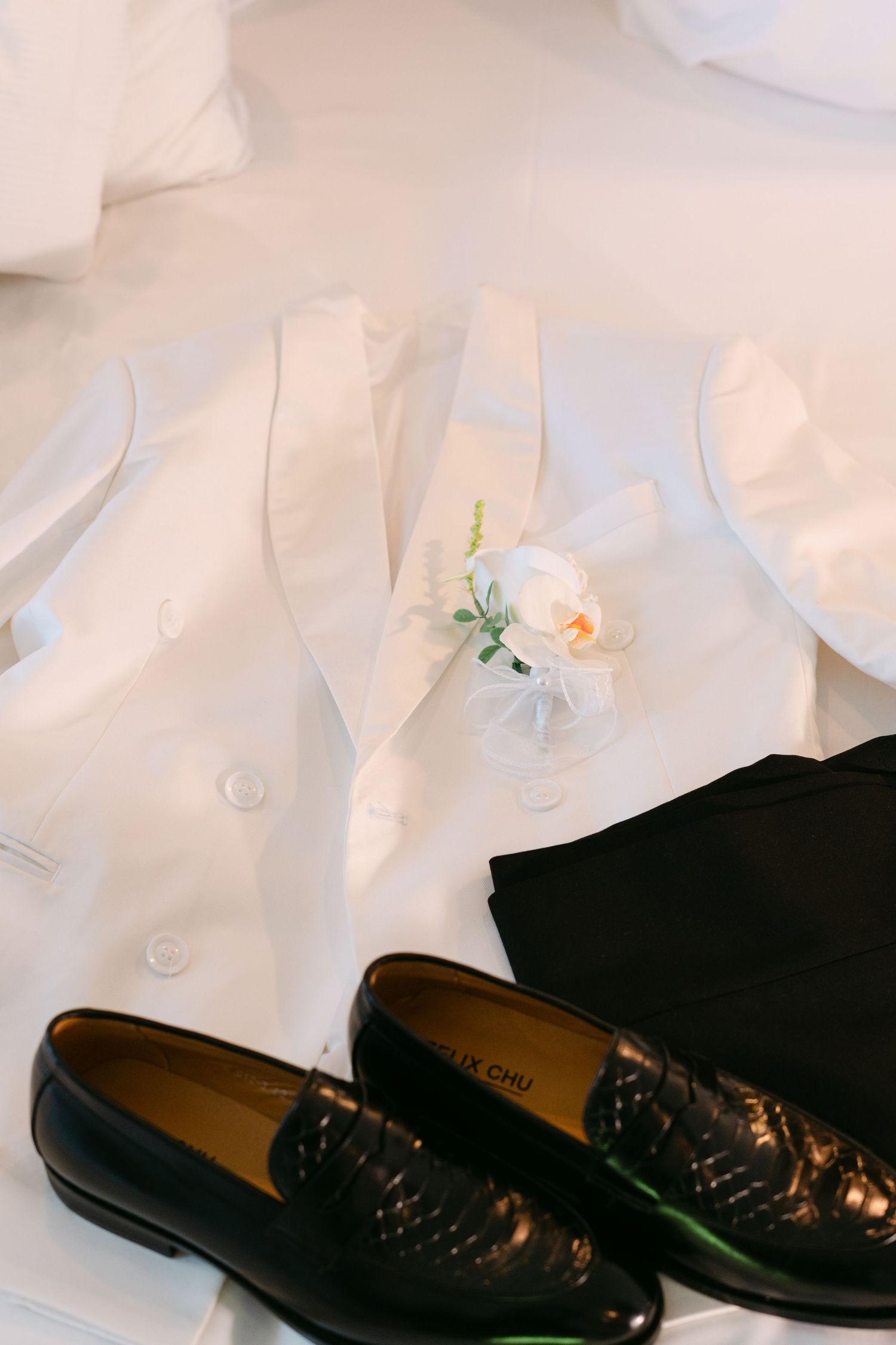 White wedding vest with floral boutonniere and black loafers displayed on white surface.