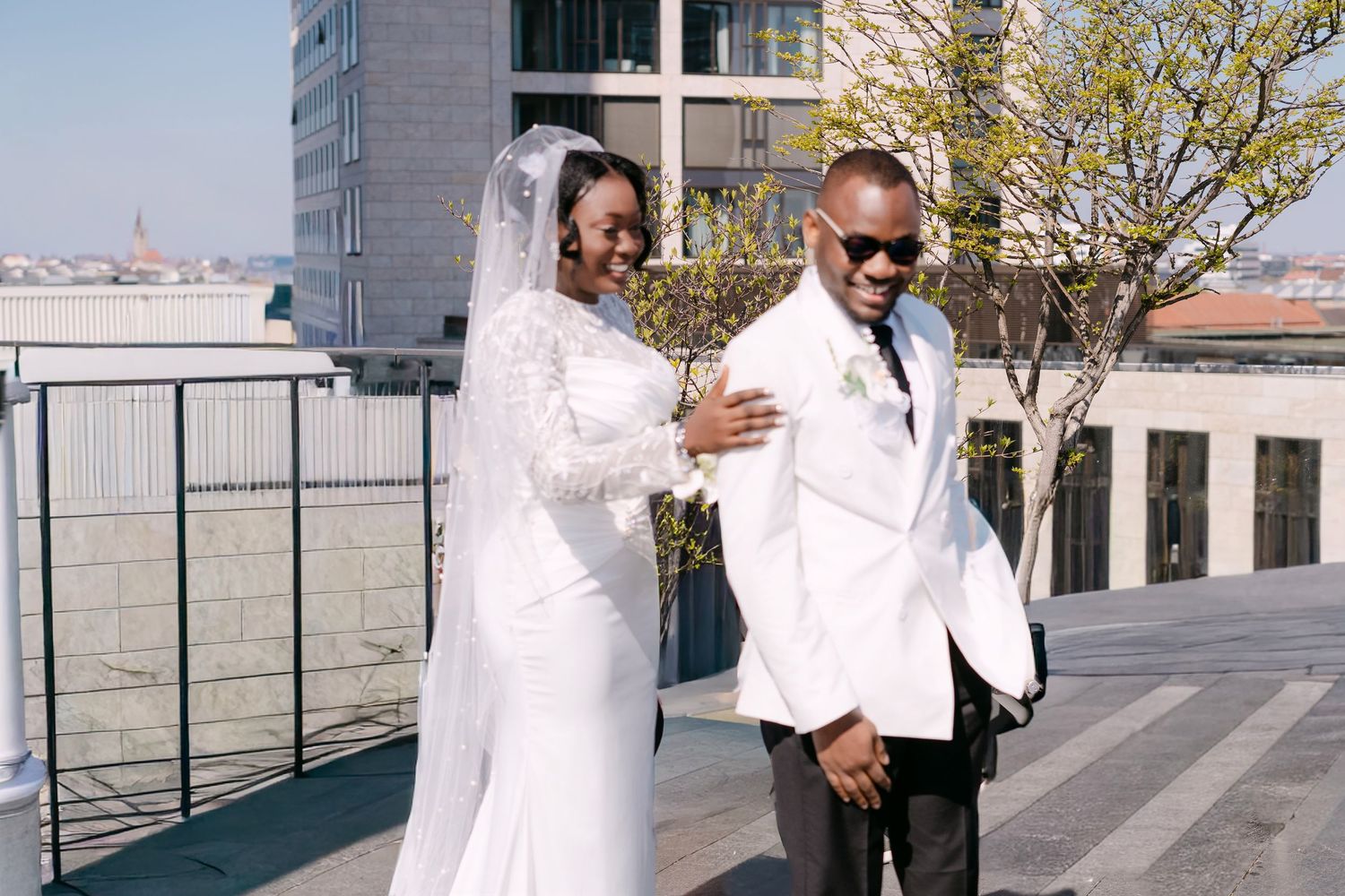 Happy African American bride and groom in white wedding attire posing on rooftop terrace.