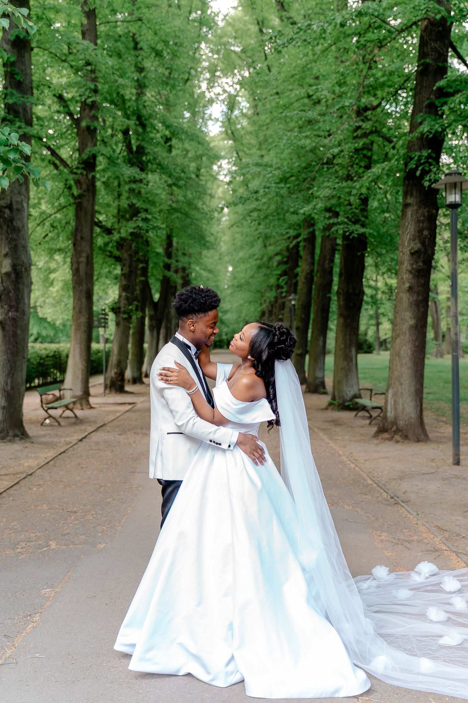 Black bride and groom share romantic moment on tree-lined path, bride wearing flowing white gown with long veil.