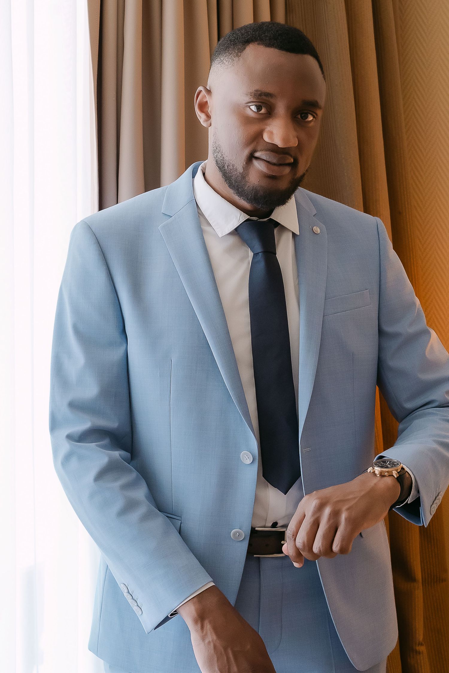 A well-dressed Black man in a light blue suit and dark tie stands confidently near a curtained window.