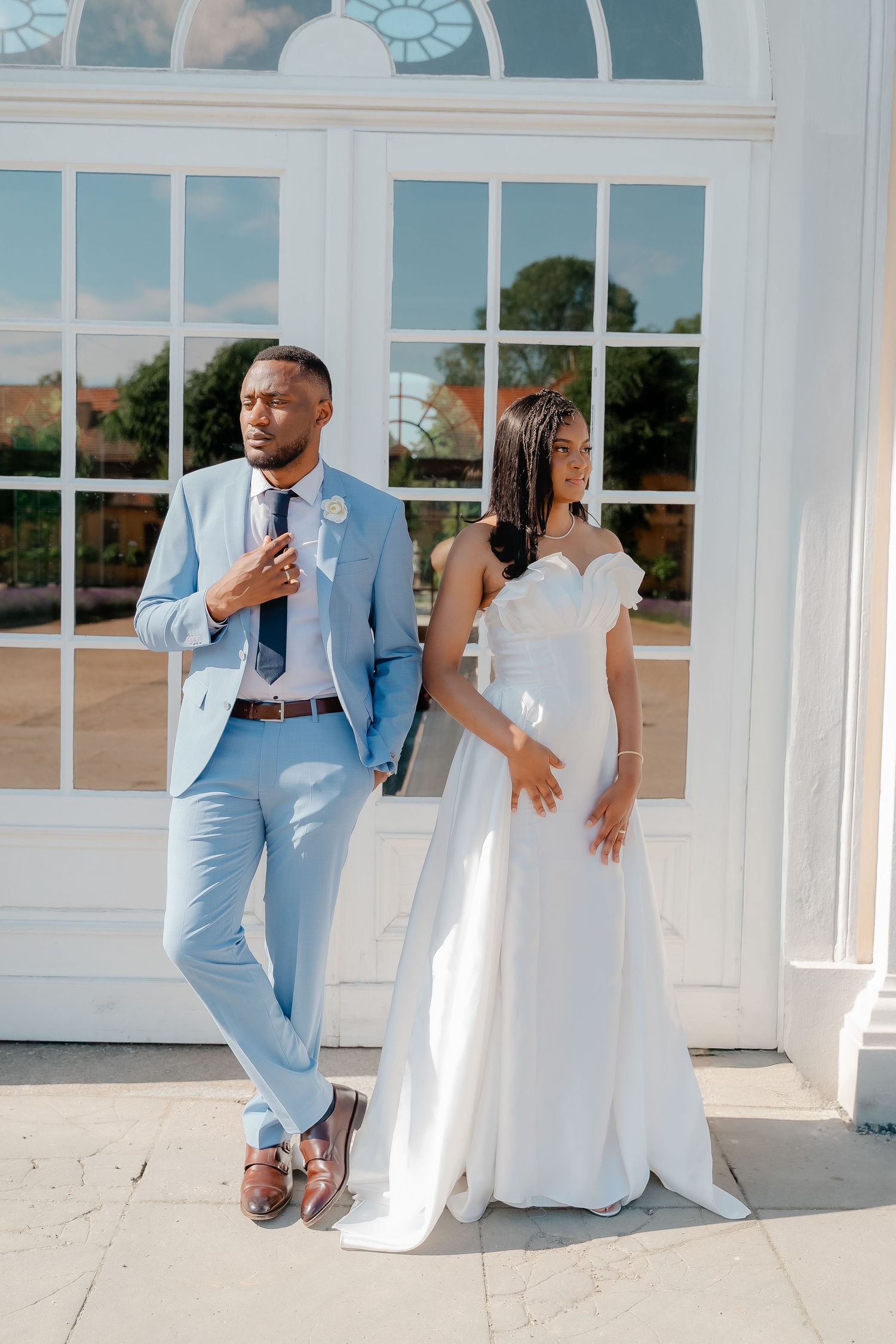 Black bride and groom in white dress and light blue suit posing before elegant white French doors.