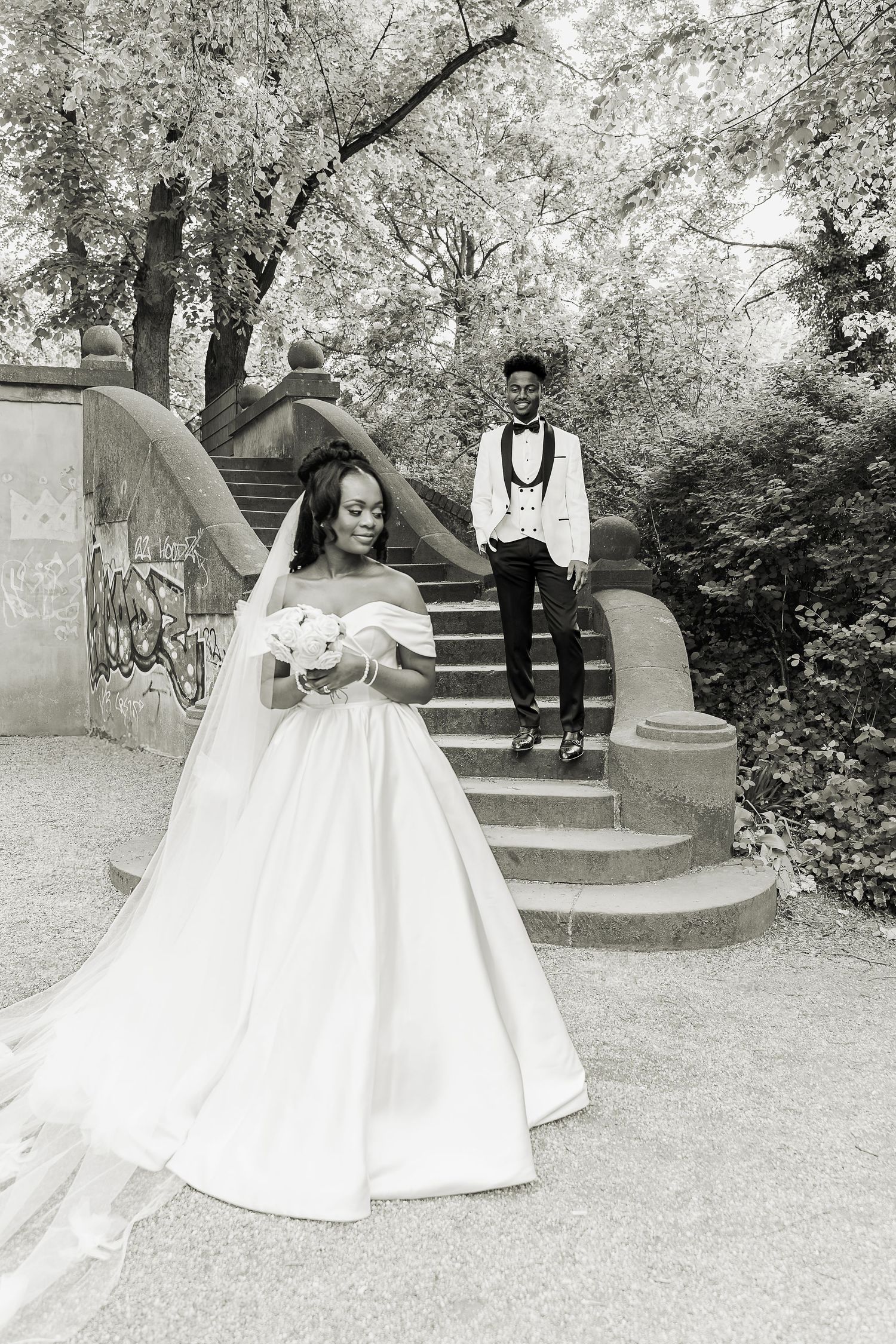 Black and white photo of Black bride in ball gown holding bouquet as groom descends stone steps in park.