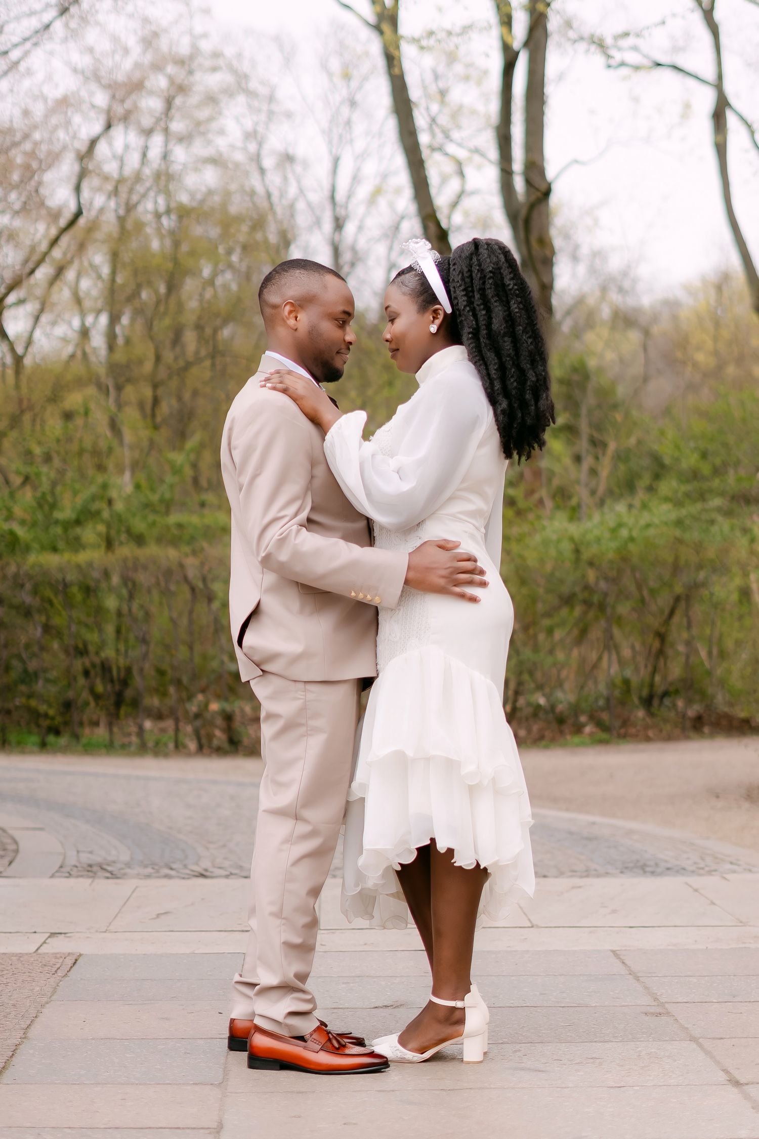 African American newlyweds embracing outdoors, groom in tan suit, bride in white ruffled midi dress.