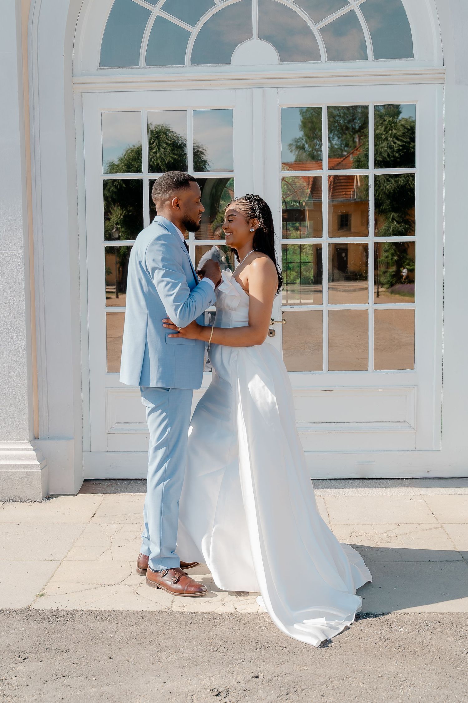 A couple in wedding attire, groom in light blue suit and bride in white gown, share a romantic moment outdoors.