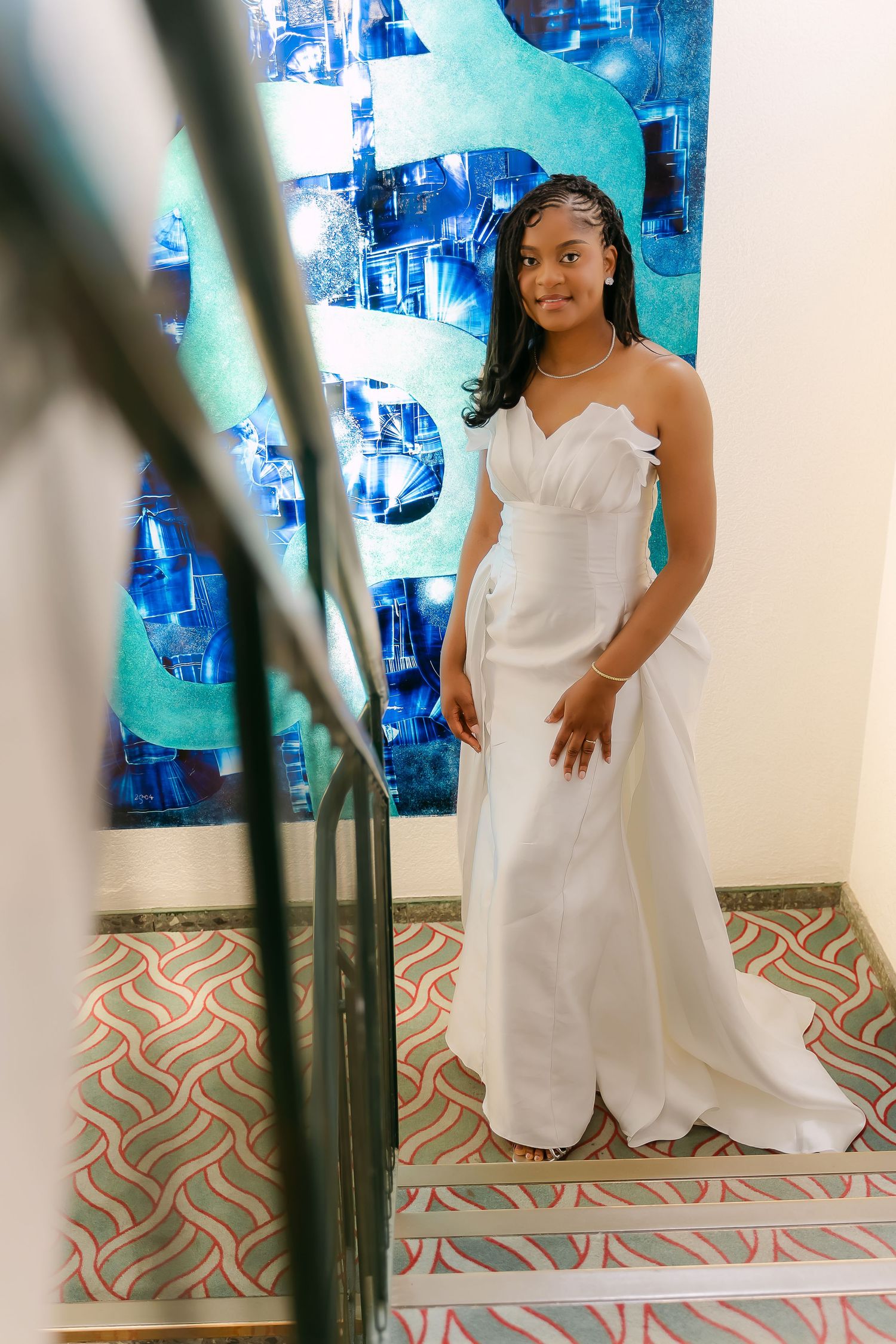 Young woman in elegant white gown poses on decorative staircase before vibrant blue abstract wall art.