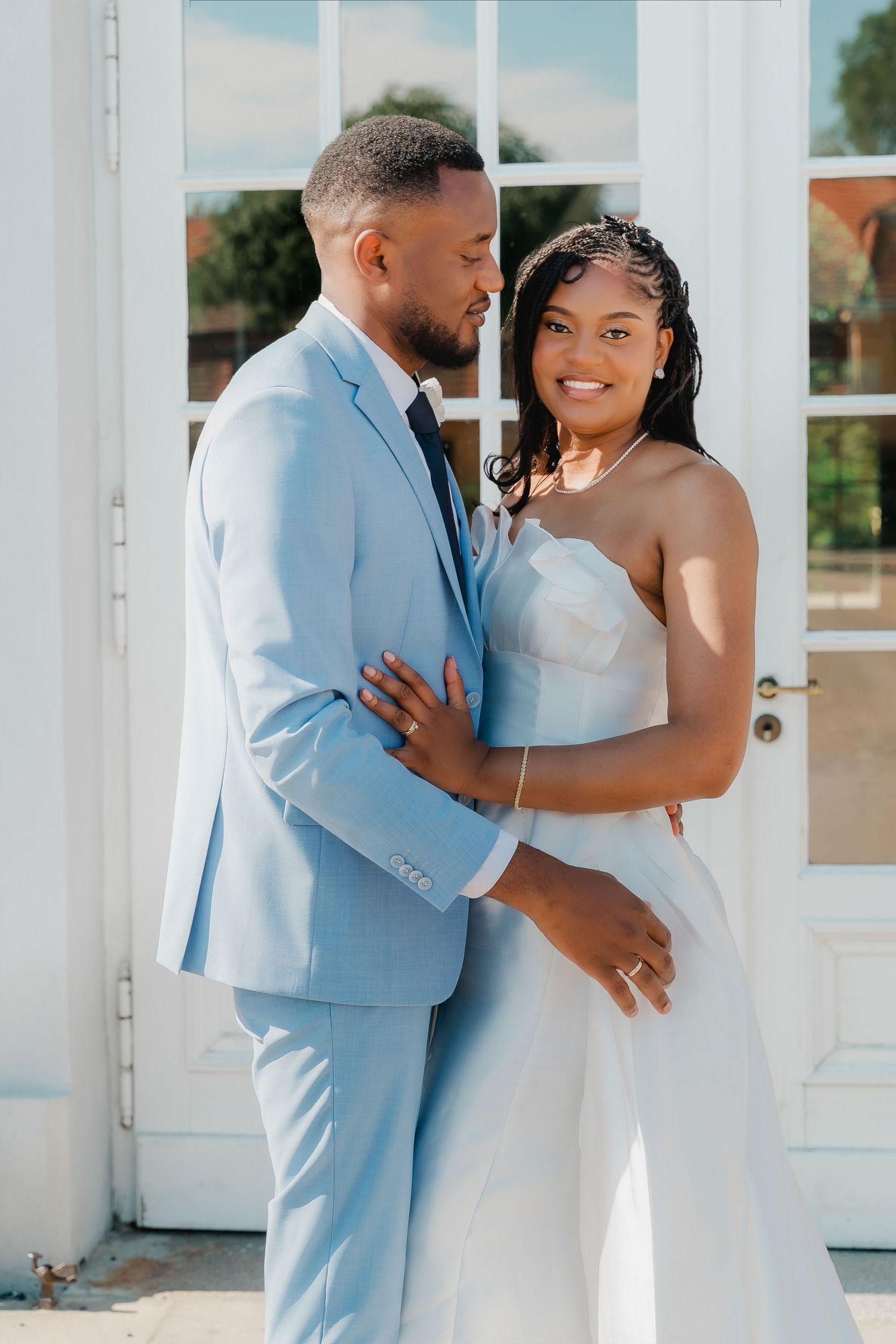 A Black couple in wedding attire embrace outdoors, groom in light blue suit, bride in white strapless gown, smiling joyfully.