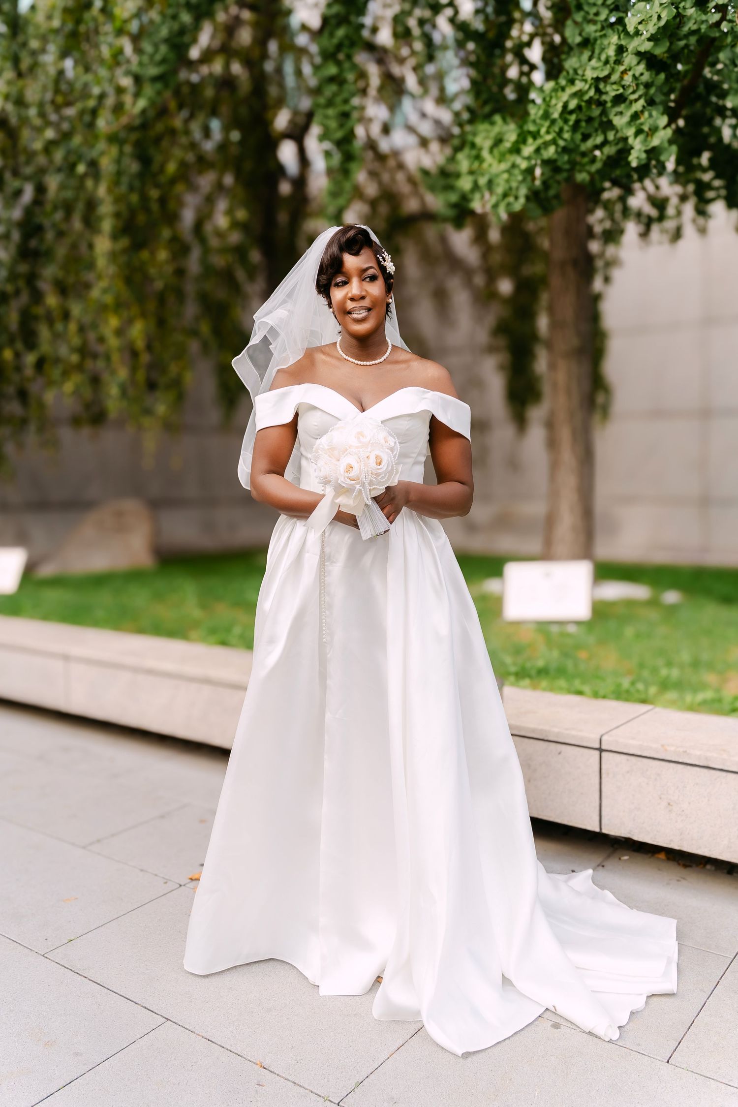 Beautiful Black bride in off-shoulder white ball gown holding bouquet outdoors on wedding day.