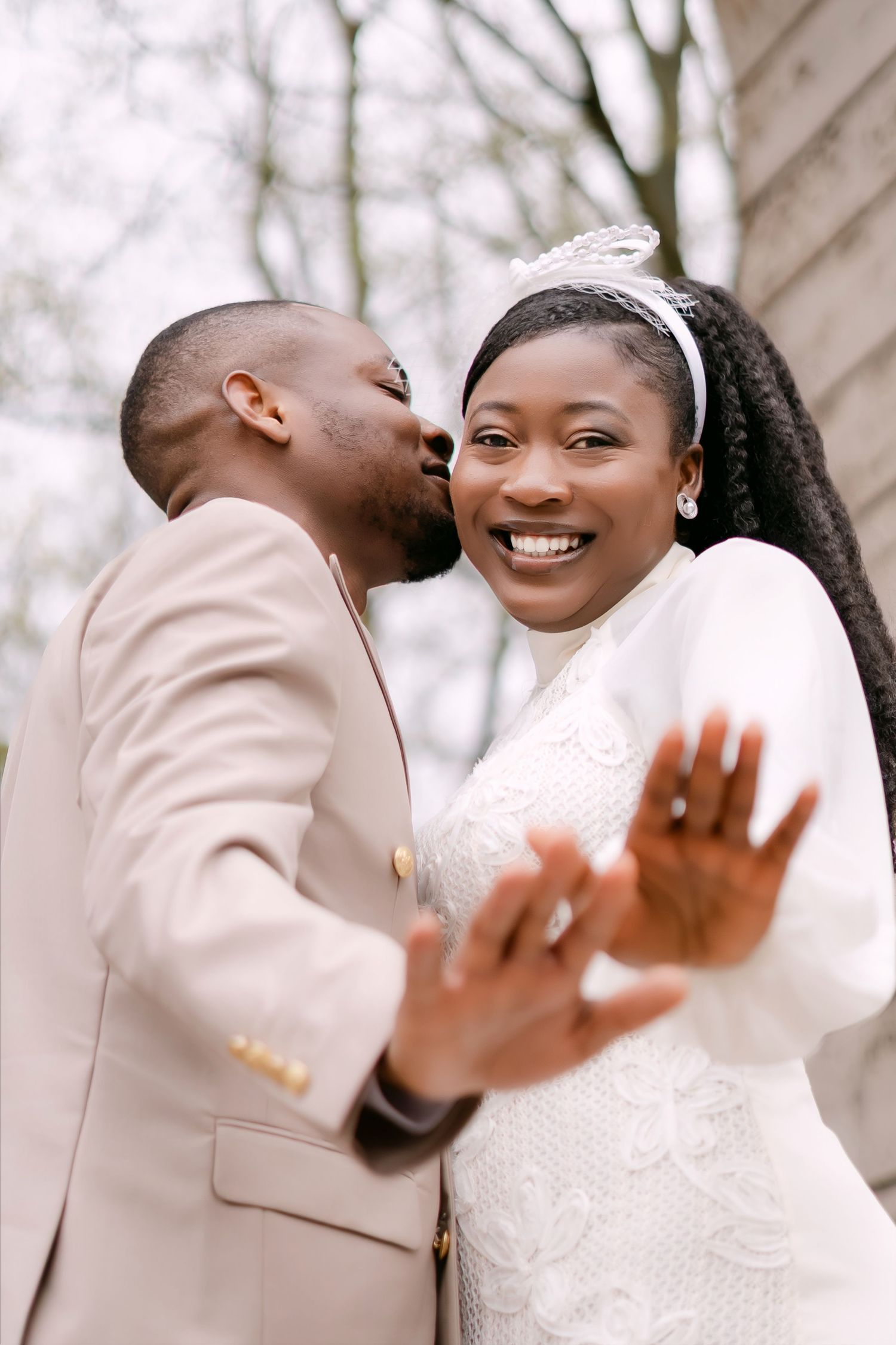 Joyful Black bride laughing as groom kisses her cheek, showing wedding rings to camera outdoors.