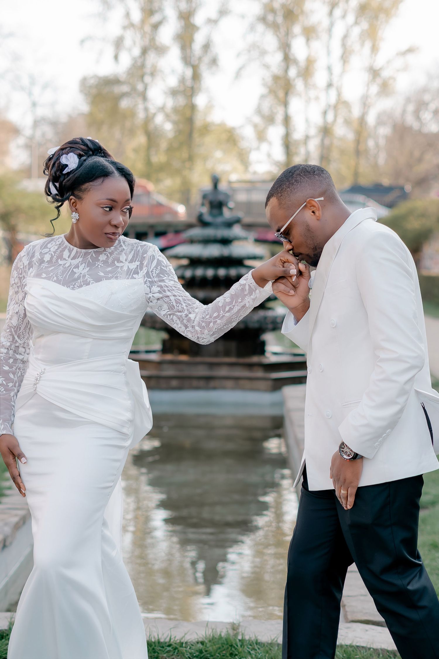Black groom in white jacket kisses bride's hand near a fountain, bride in elegant lace wedding gown.