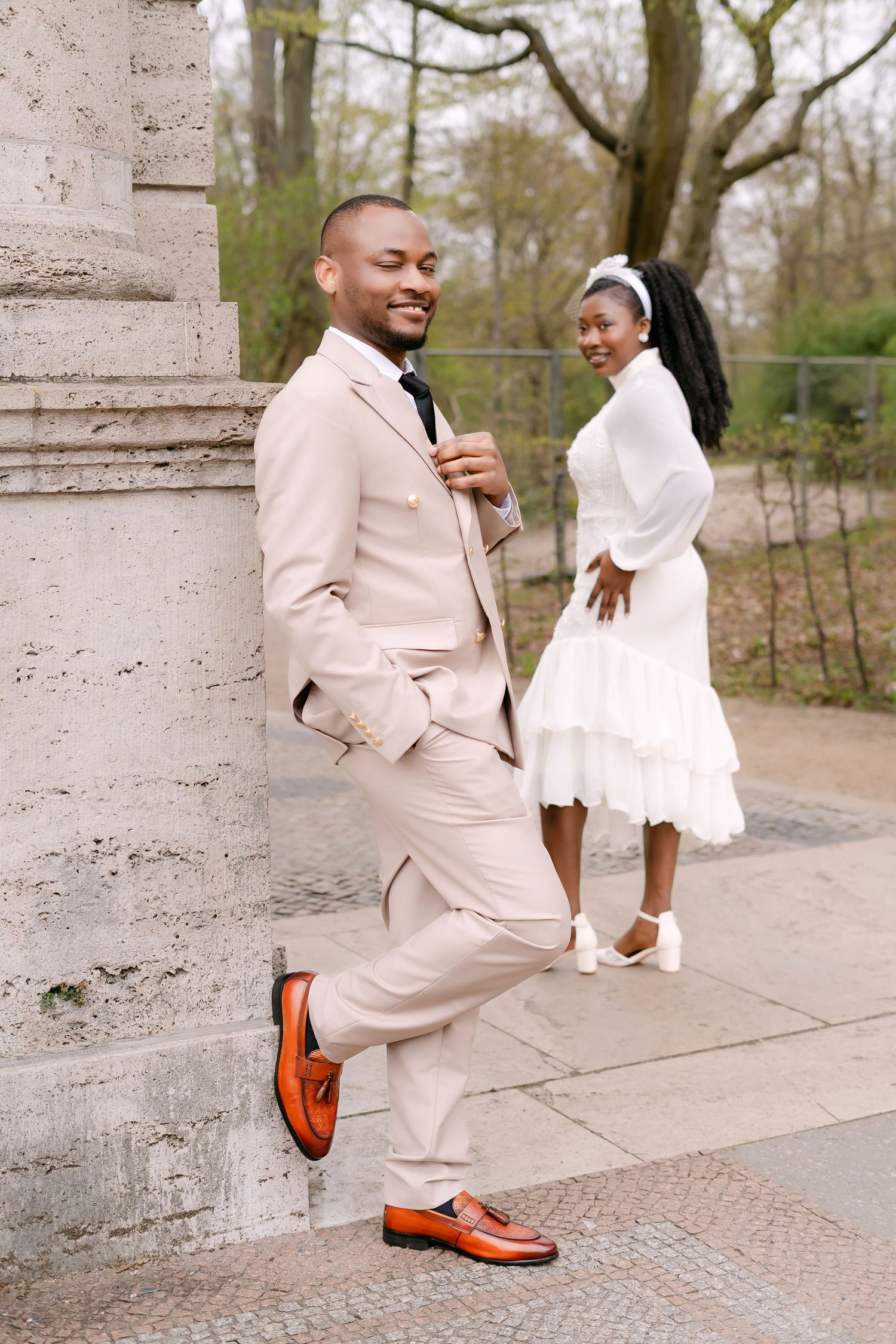 A groom in a beige suit smiles while leaning on a stone column as his bride stands behind him in white.
