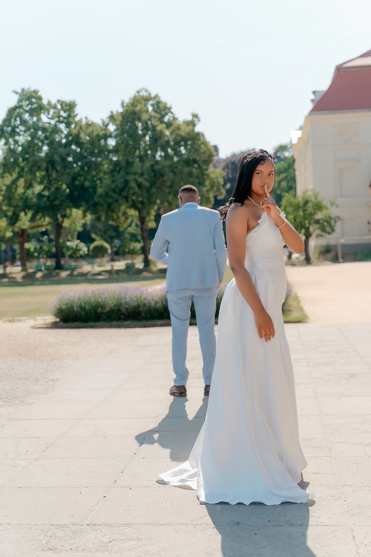 Black bride in white strapless gown smiling as groom walks ahead in a sunny outdoor garden setting.