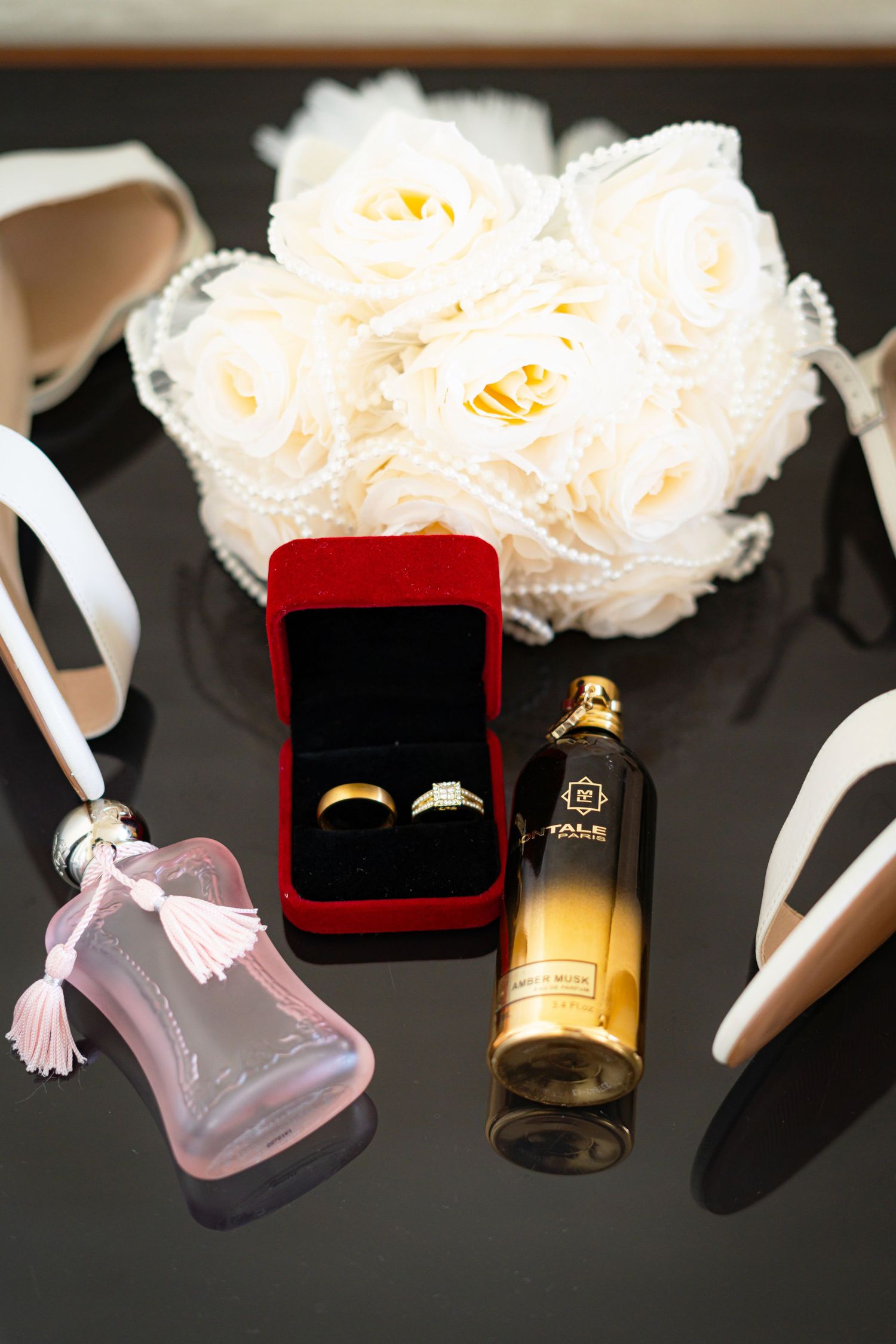 Wedding flat lay featuring gold rings in red box, perfume bottles, white rose bouquet, and heels.
