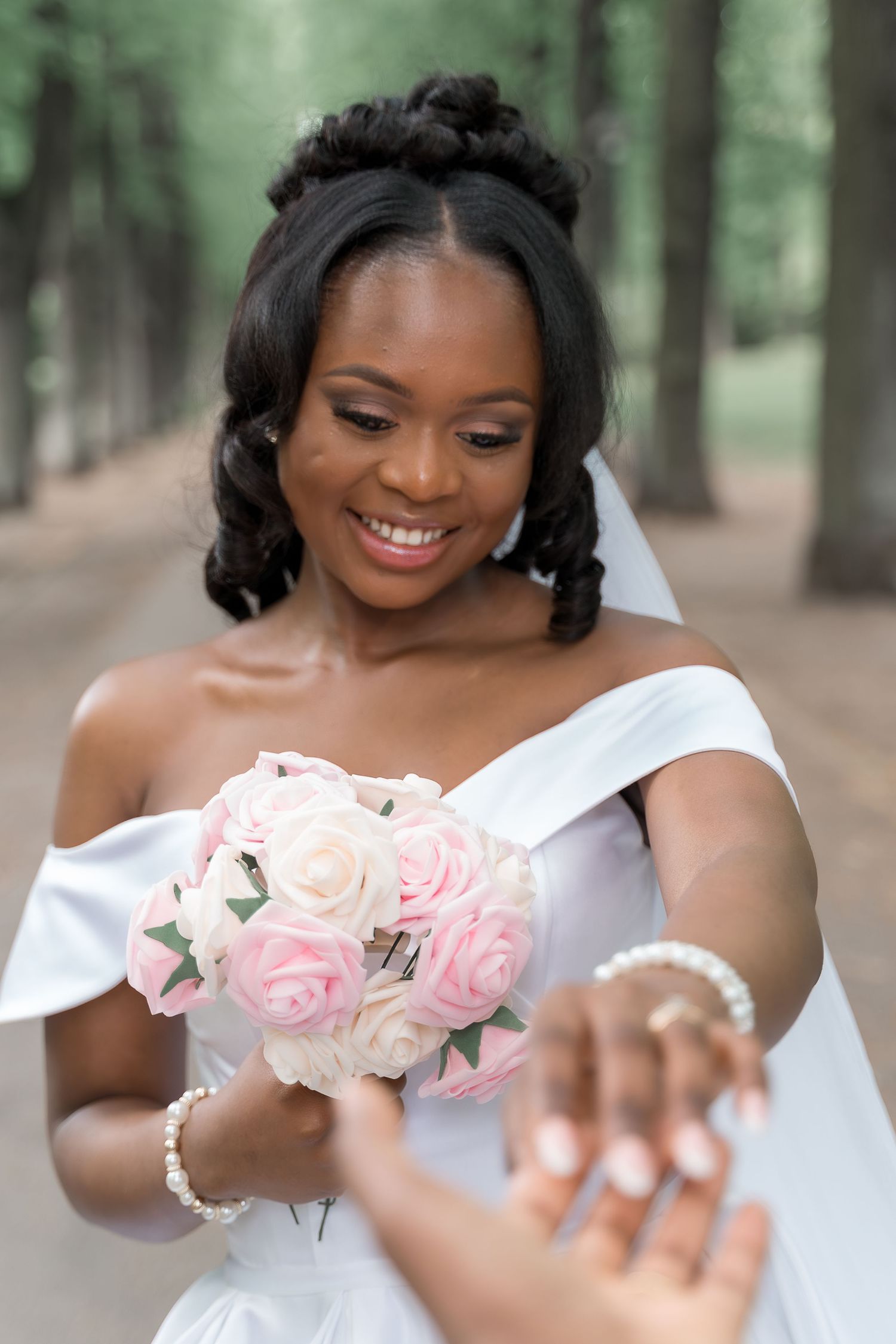 Smiling Black bride in white off-shoulder gown holds pink rose bouquet outdoors in a tree-lined path.
