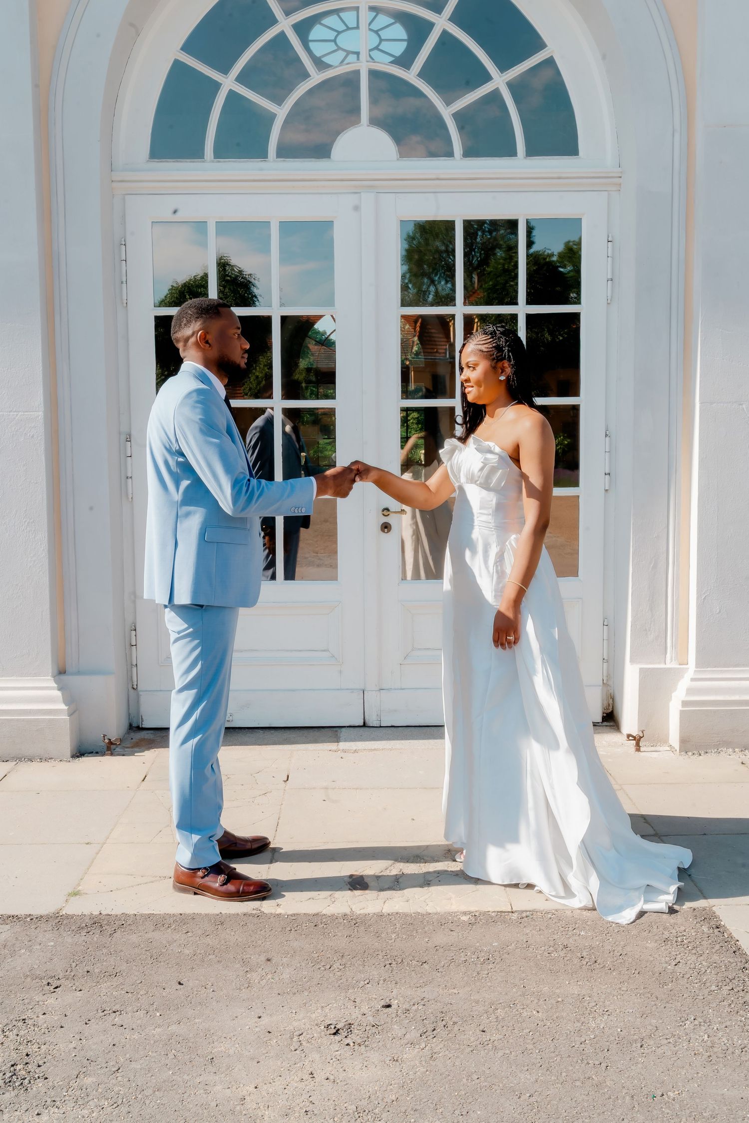 A bride and groom hold hands in front of a white arched door, she in a white gown, he in a light blue suit.