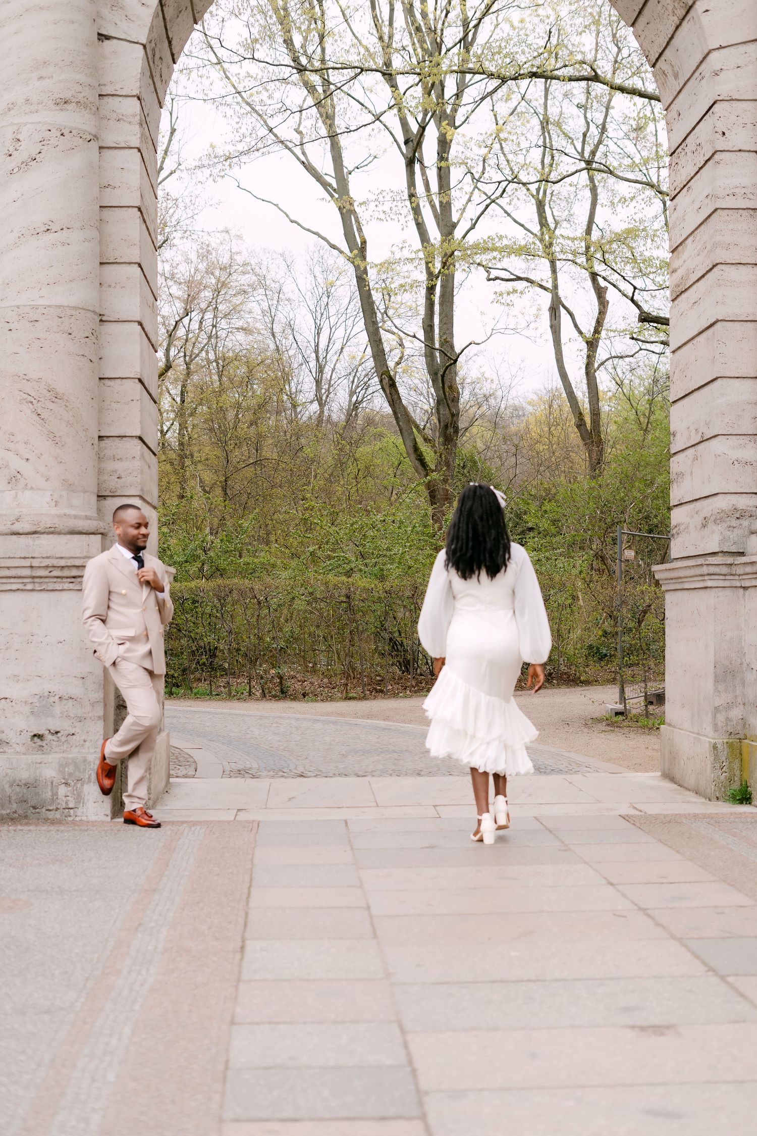 A groom in a beige suit leans against a stone column as his bride in a white dress walks toward him.