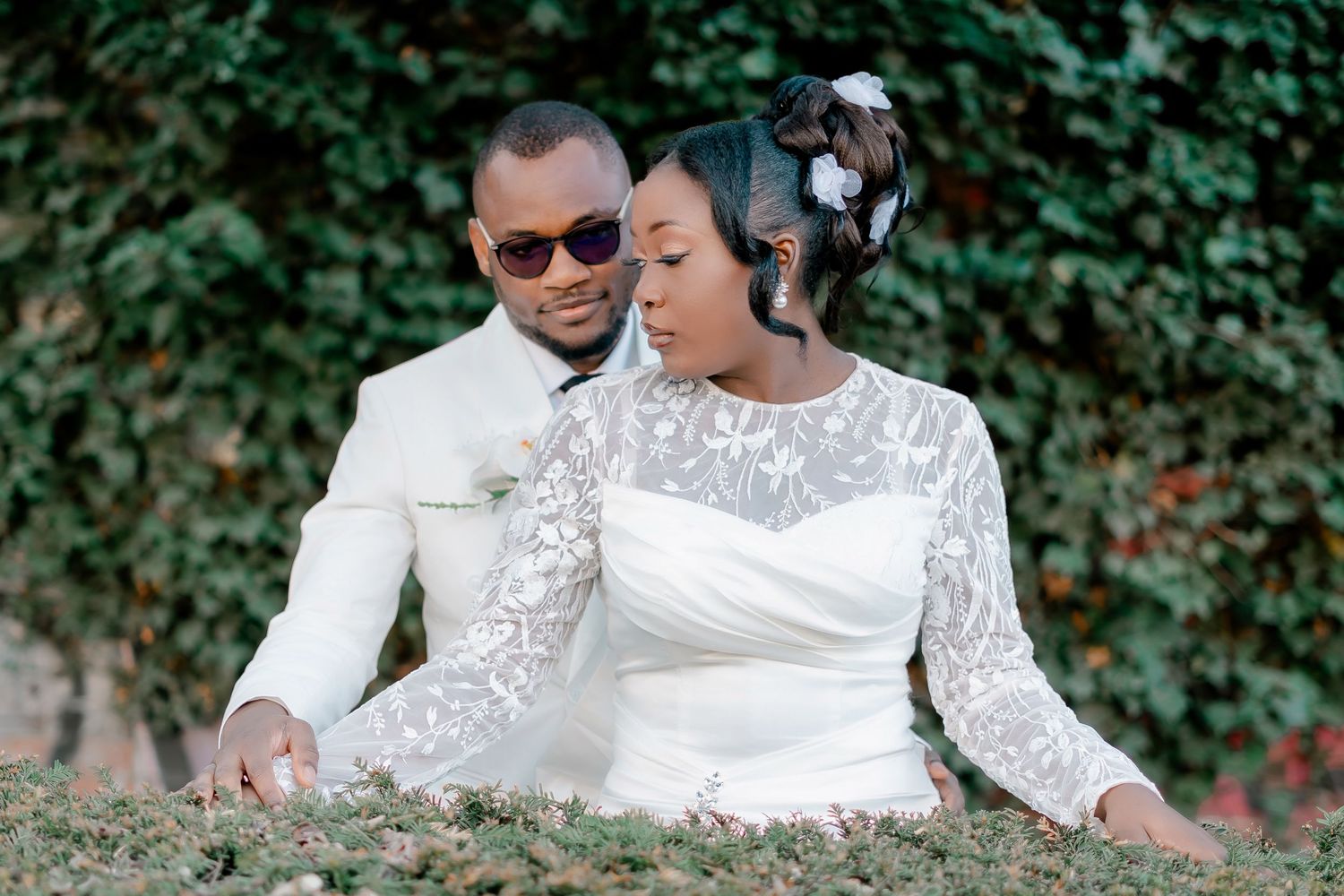 Black bride and groom in white wedding attire posing together against a lush ivy-covered wall.