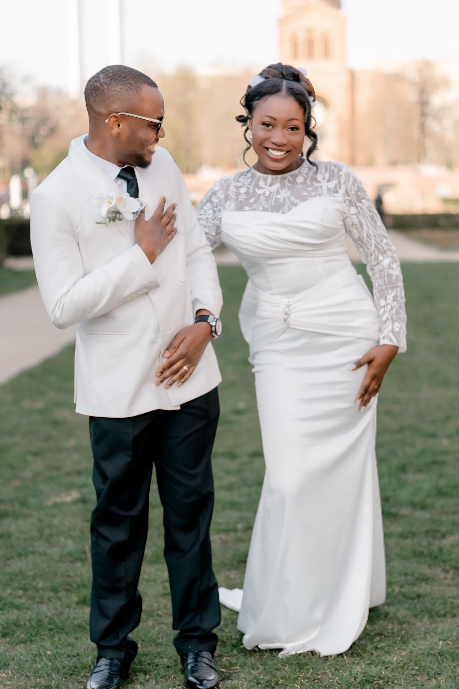 Smiling bride in lace wedding gown with groom in white blazer on green lawn.