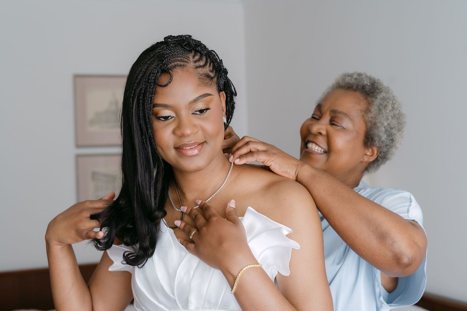 Elderly woman lovingly placing a necklace on a smiling Black bride during wedding preparations.