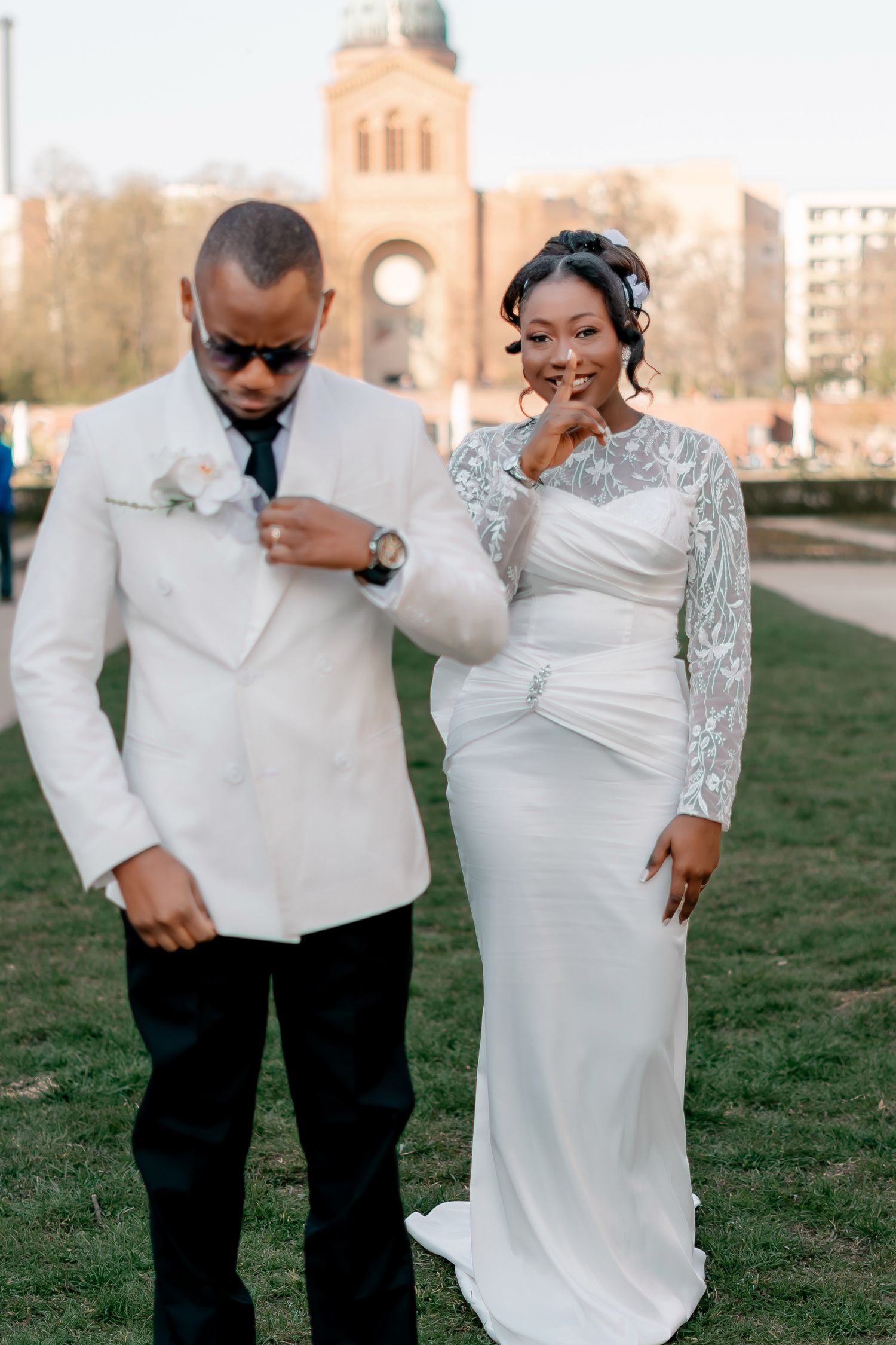 Stylish groom in white suit with sunglasses and bride in lace wedding dress outdoors.