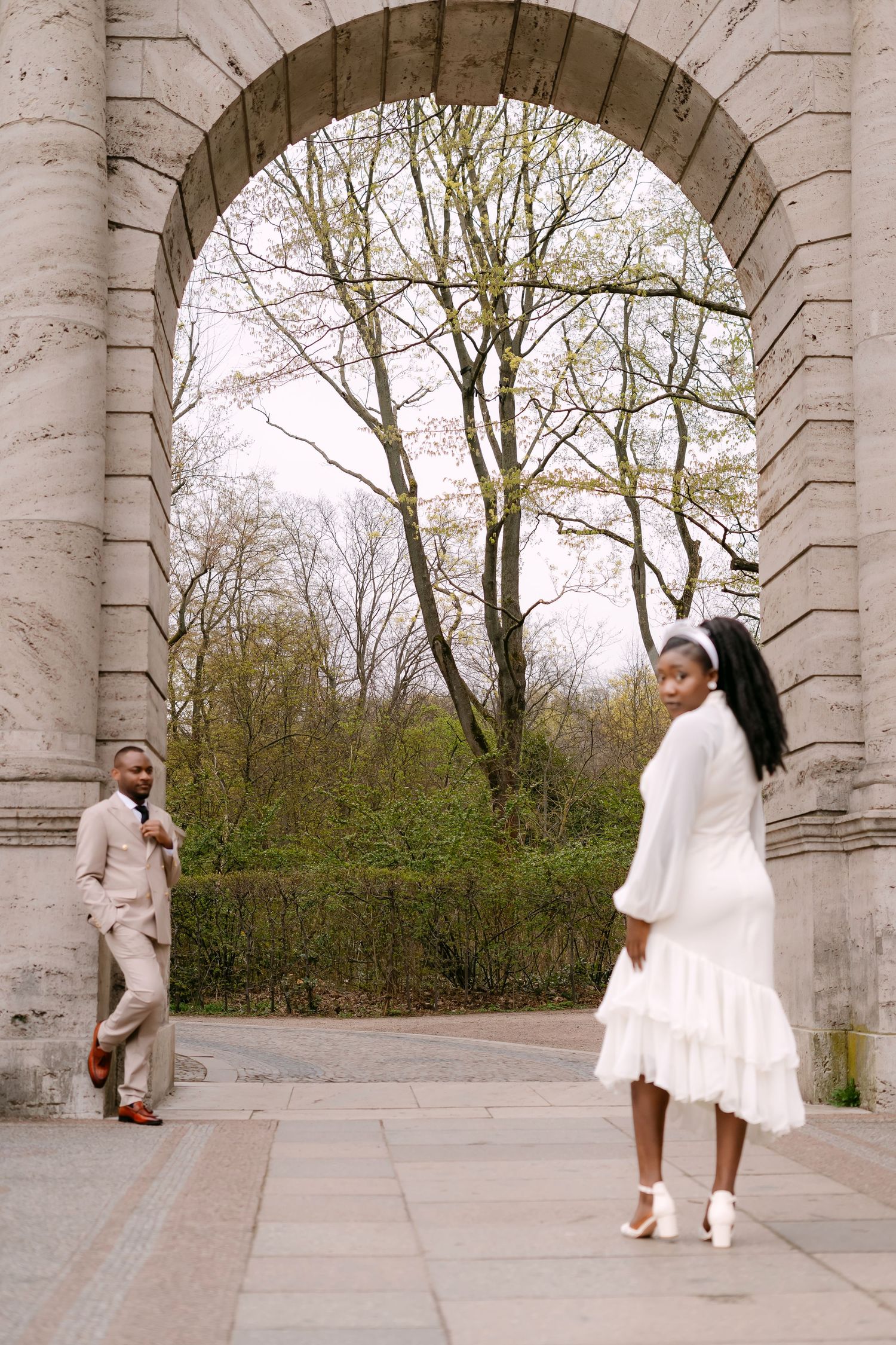 A bride in a white dress looks back at her groom in a beige suit standing under a grand stone archway.